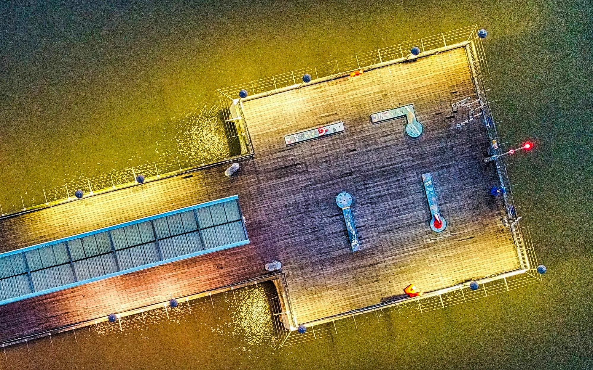 Overhead view of a wooden pier extending over water, with game tables and benches on it, illuminated at night.