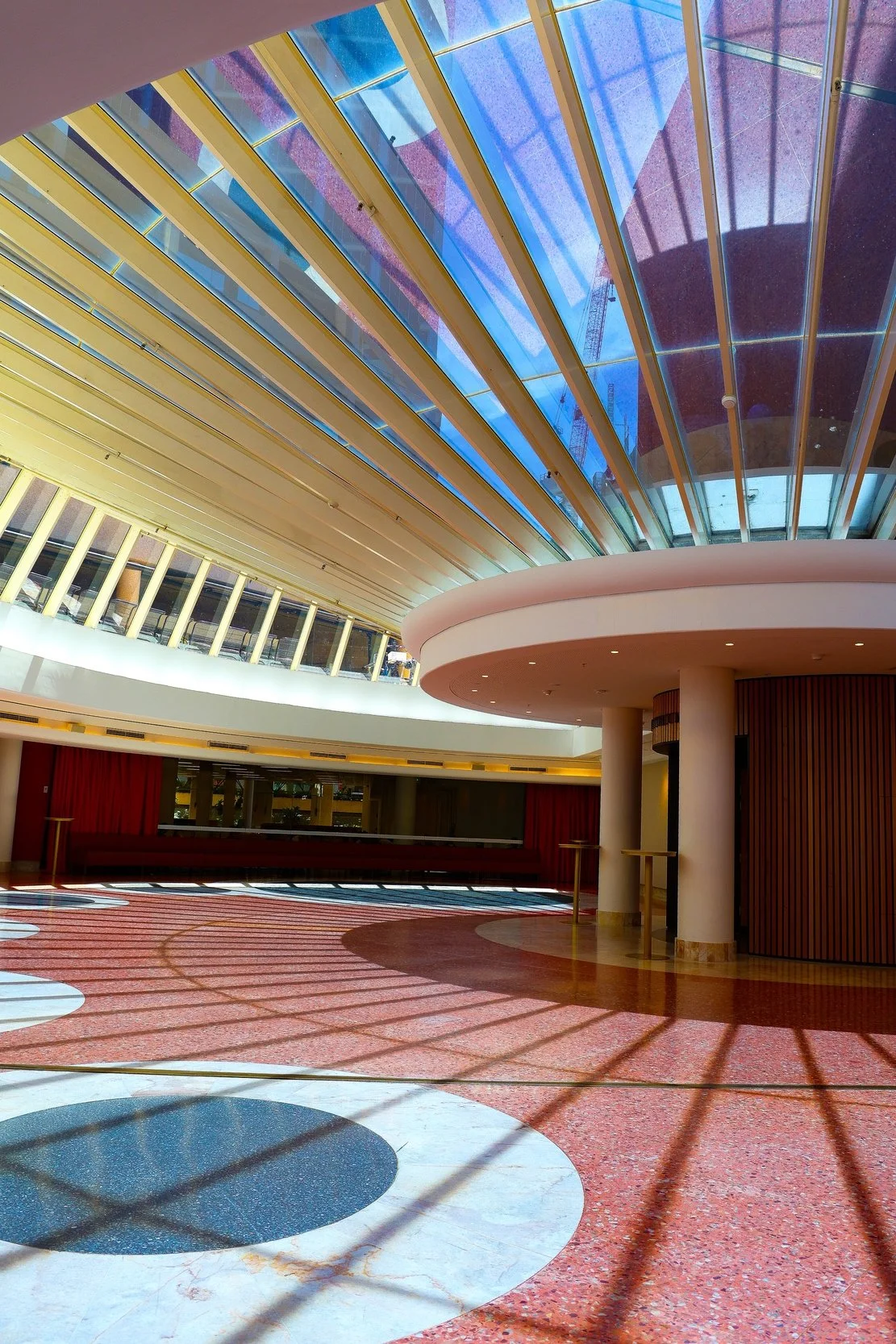 Interior view of a modern building with a colorful patterned red and white floor, large glass ceiling with yellow beams, and a reflective curved wall with red curtains.
