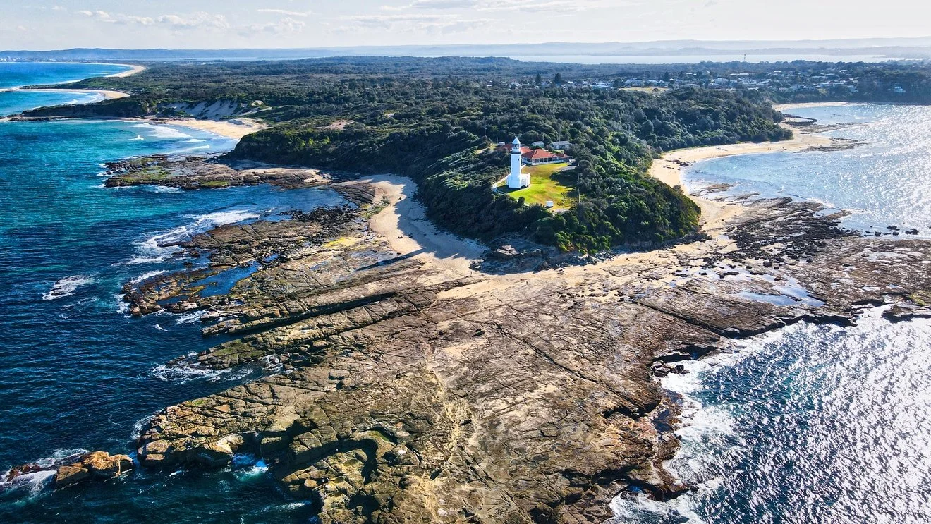 Aerial view of a coastal landscape with rocky shoreline, sandy beaches, a green wooded area, and a white lighthouse on a hilltop overlooking the ocean.