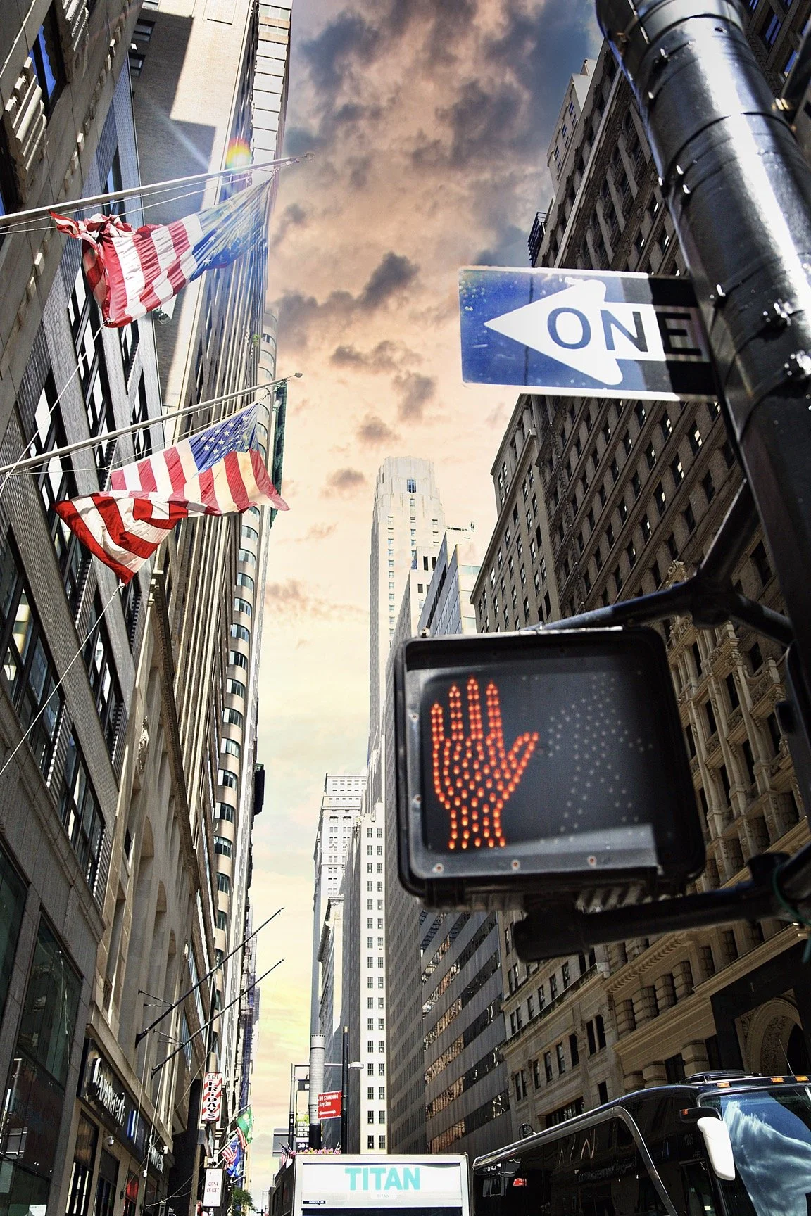City street scene with tall buildings, American flags, a traffic signal with a red hand, and a one-way sign pointing to the left at sunset.
