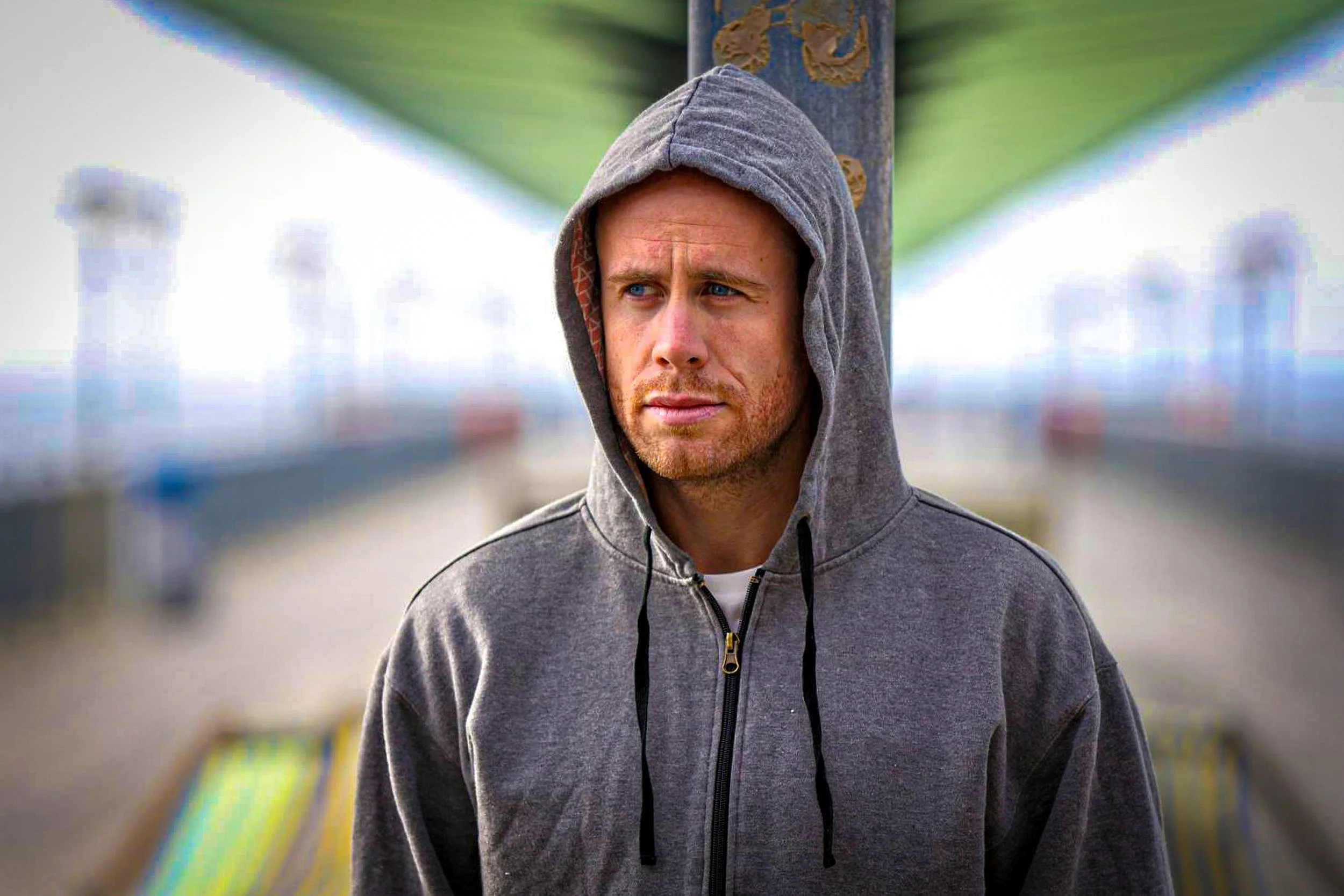 A man with a contemplative expression, wearing a gray hoodie with the hood up, standing outdoors under a structure with a blurred background.