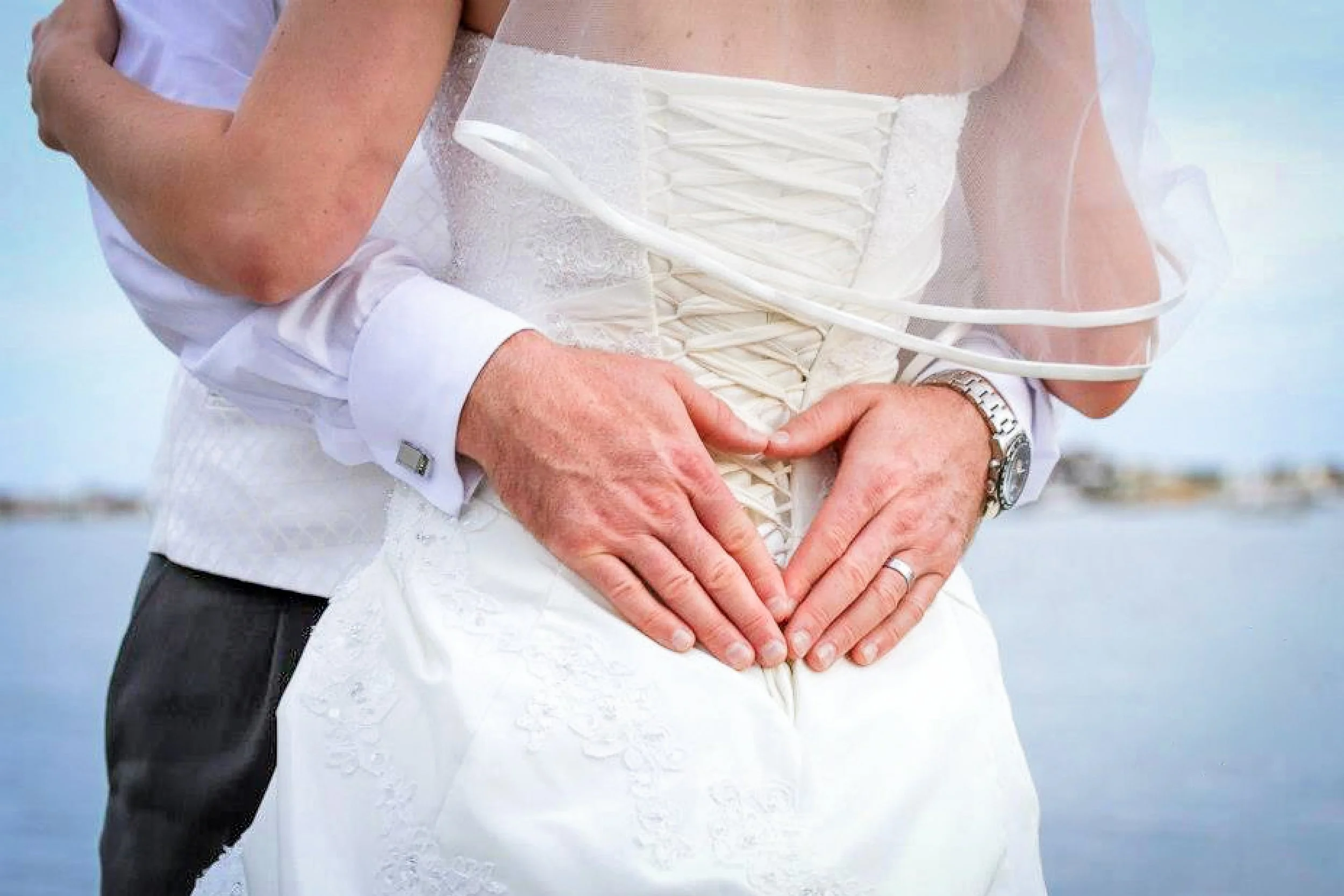 A couple in wedding attire making a heart shape with their hands at the back of a person, outside near a body of water.