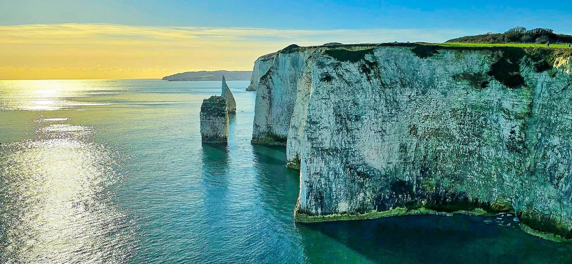 Cliffs at Étretat on the coast of Normandy, France, with white chalk formations and a small patch of grassy area at the top, under a blue sky with a few clouds, and a calm sea reflecting sunlight.