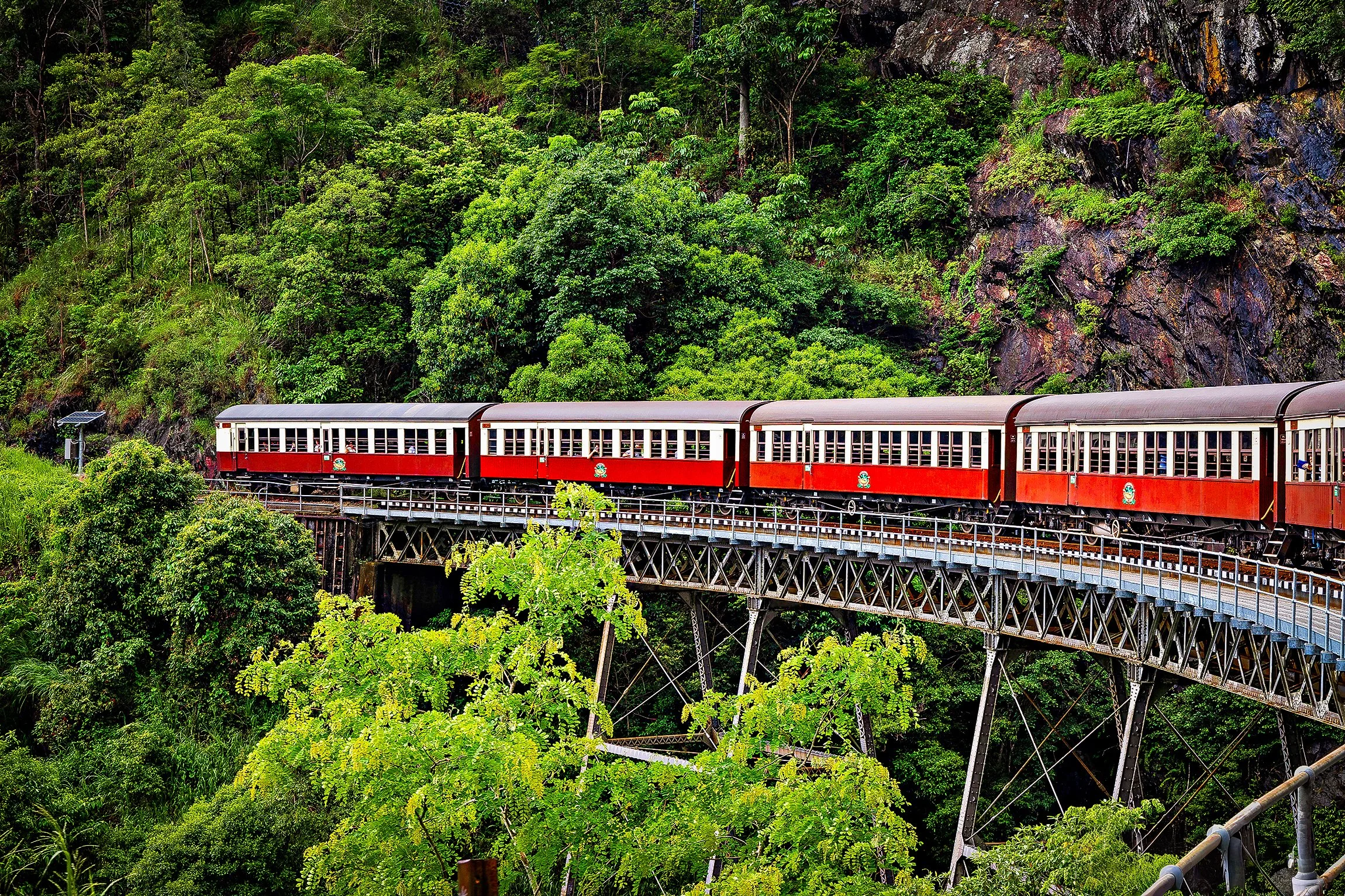 A red and white train traveling on a bridge through a lush green forest with rocky cliffs in the background.