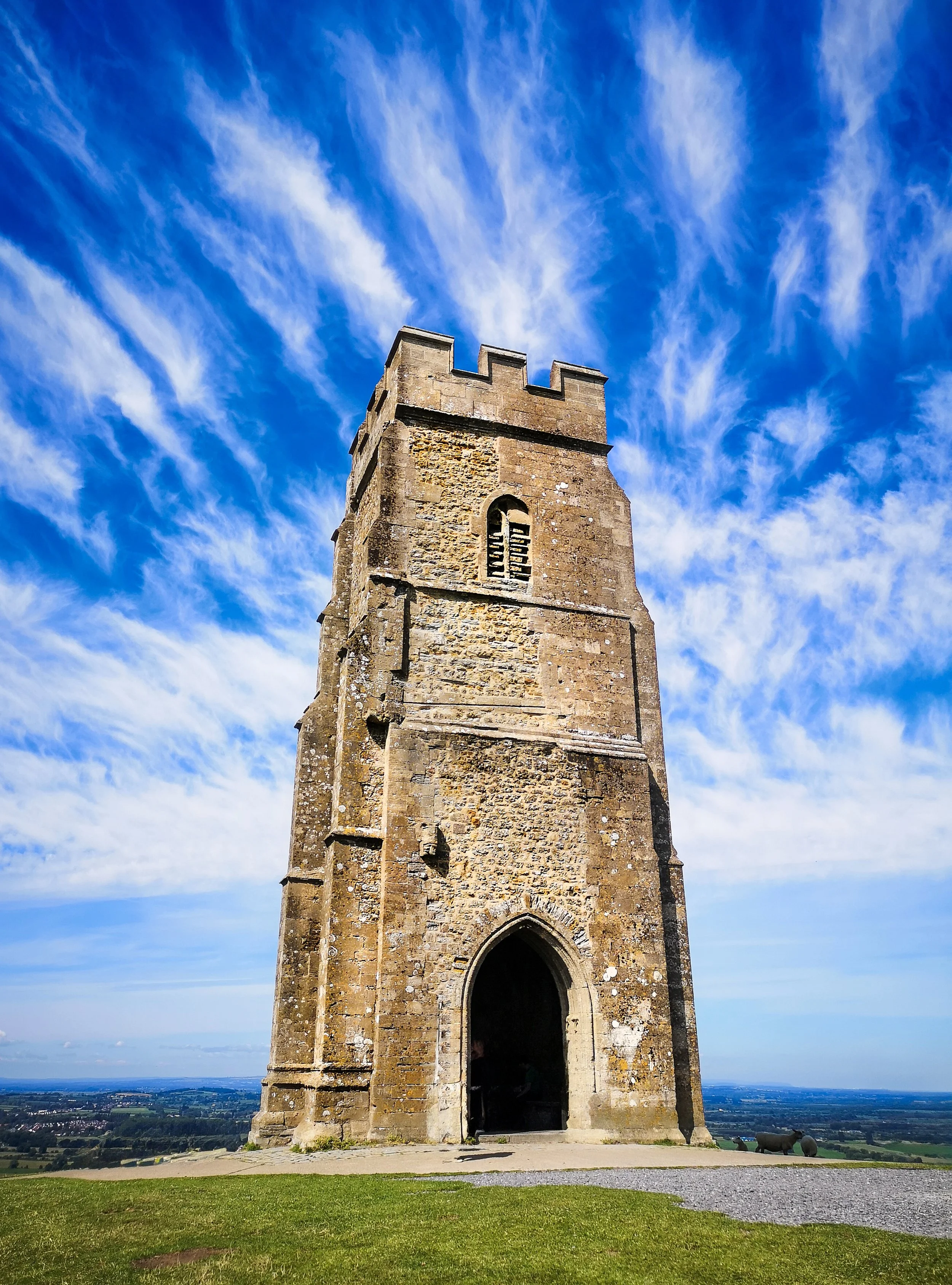 A tall, ancient stone tower with a crenellated top standing on a grassy landscape under a blue sky with wispy white clouds.