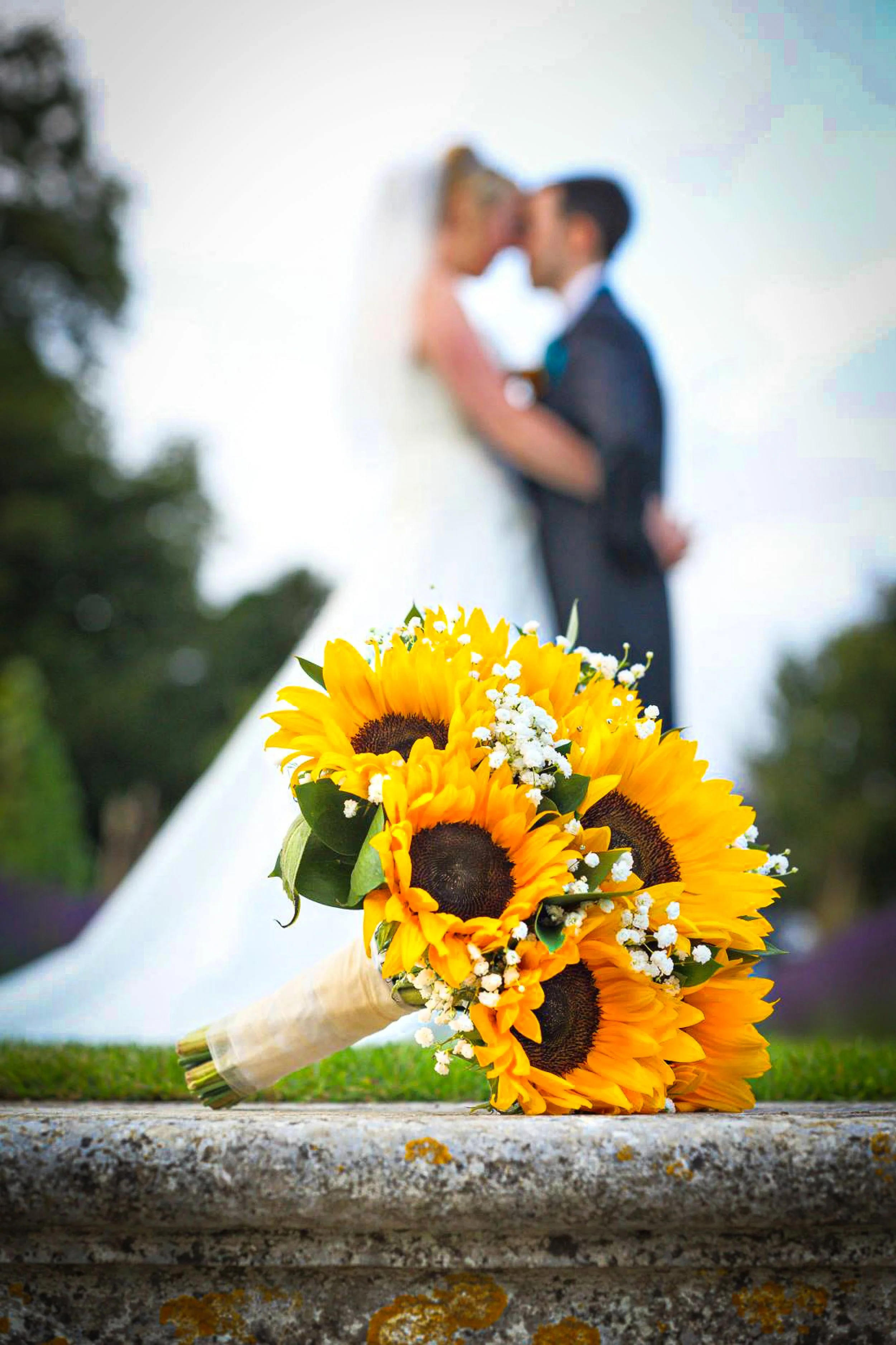 A bridal bouquet of sunflowers with white baby's breath rests on a stone surface, with a blurred bride and groom standing closely together in the background.