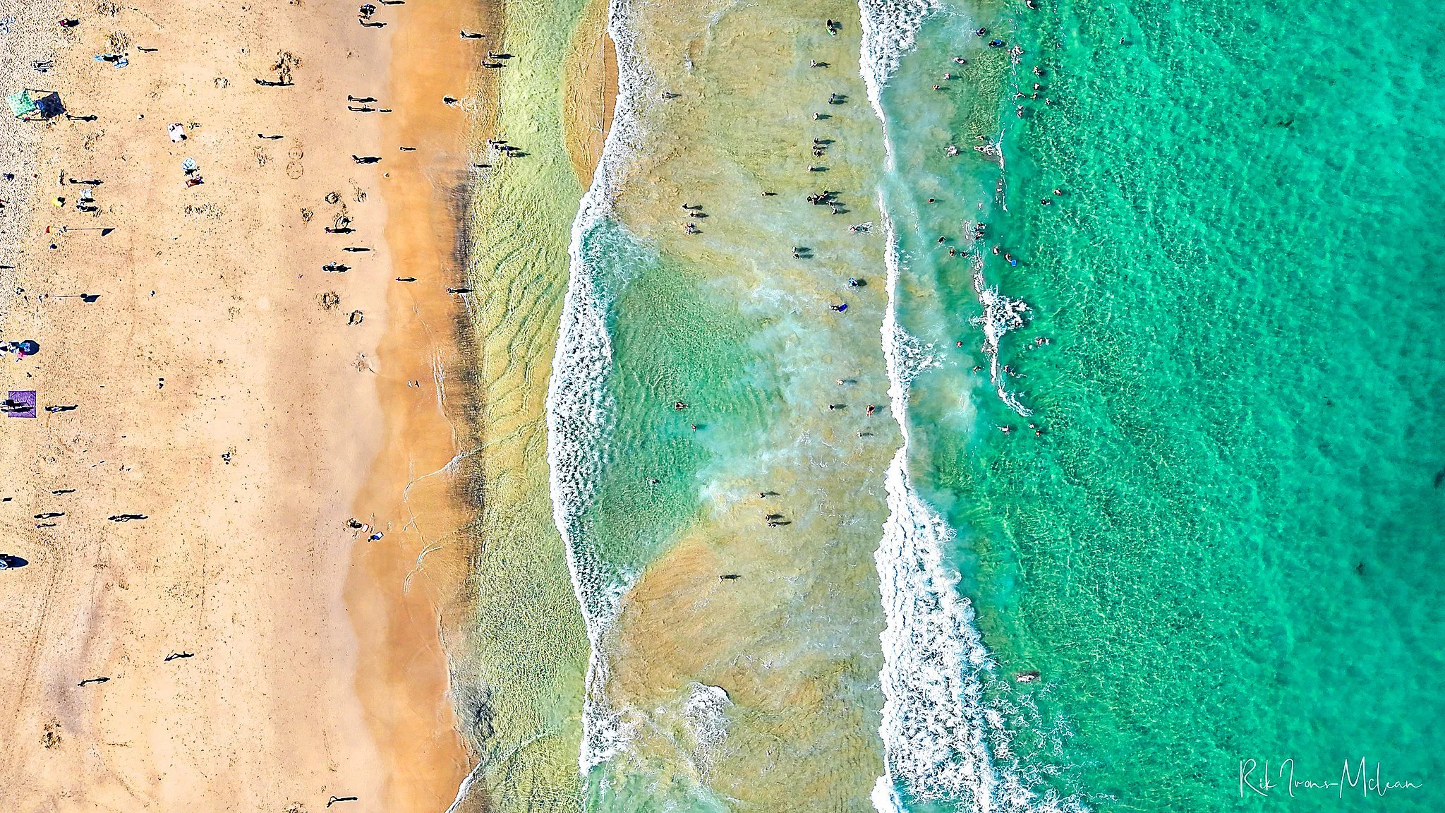 Aerial view of a sandy beach with people walking and sunbathing, and the ocean with waves and swimmers.