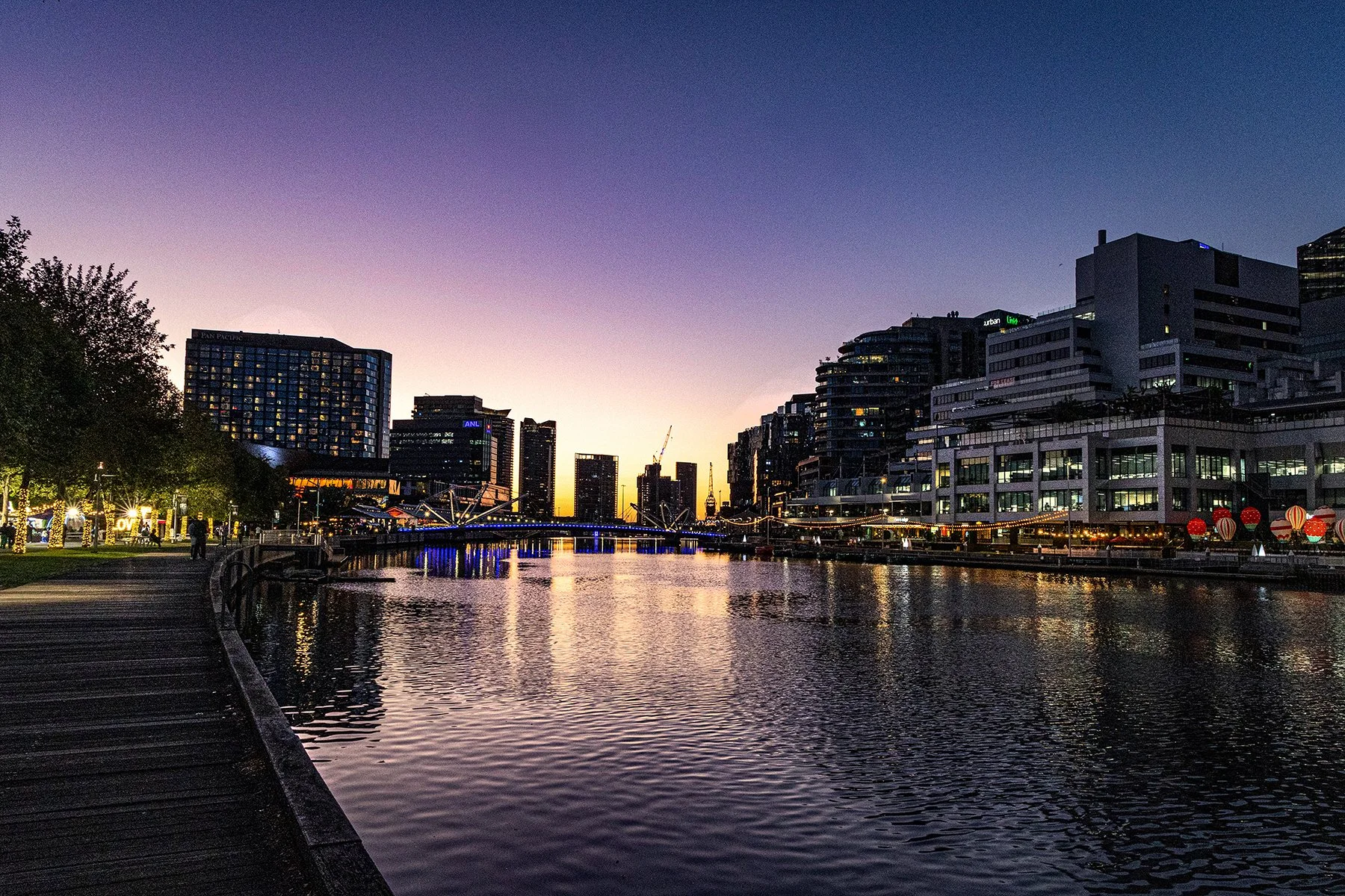 A cityscape during sunset with water reflecting the buildings and sky, featuring modern high-rise buildings, a bridge with lights, and a walkway with trees on the left.