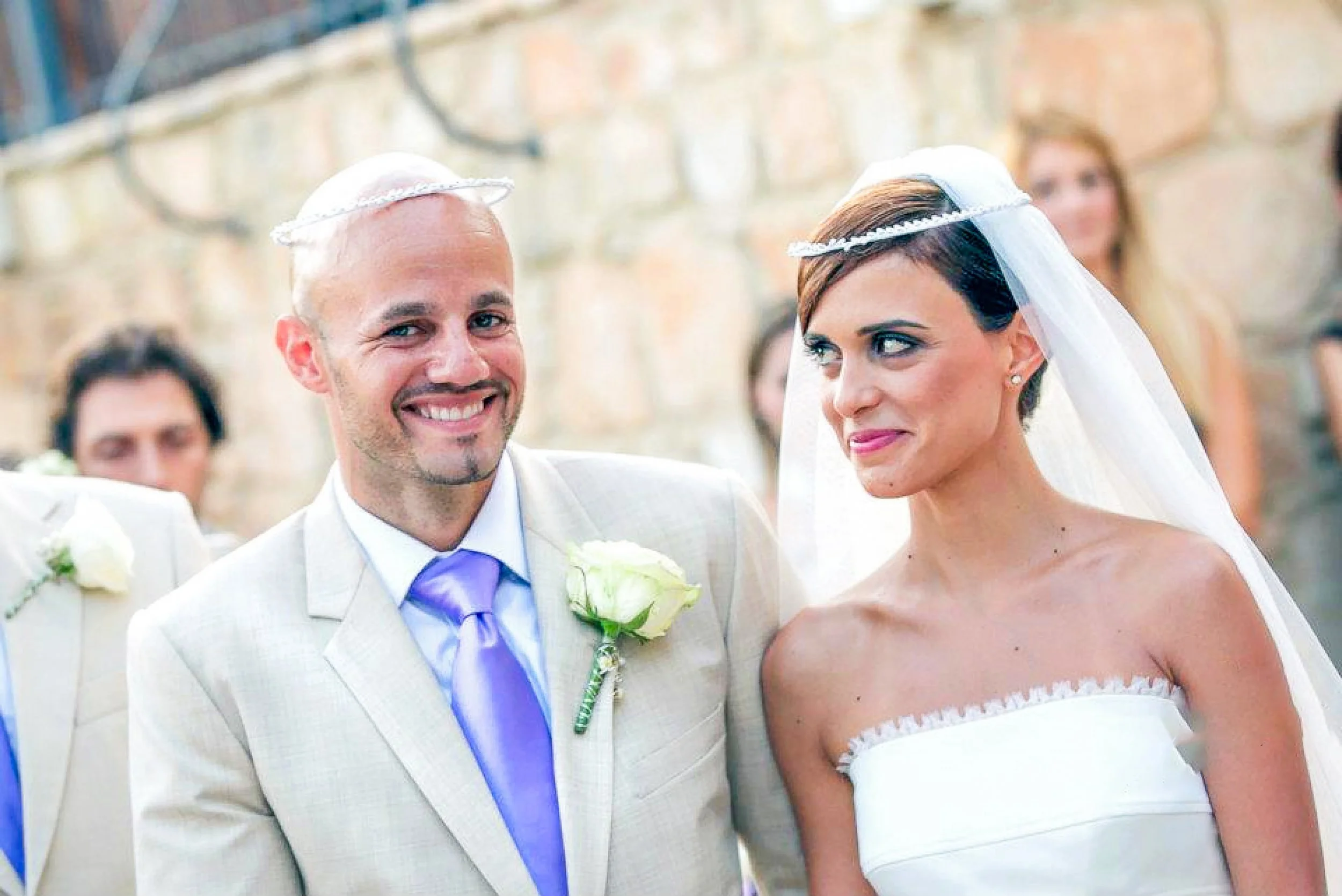 Wedding couple, man in beige suit with purple tie and woman in white strapless wedding dress, standing outdoors during ceremony with friends in background.