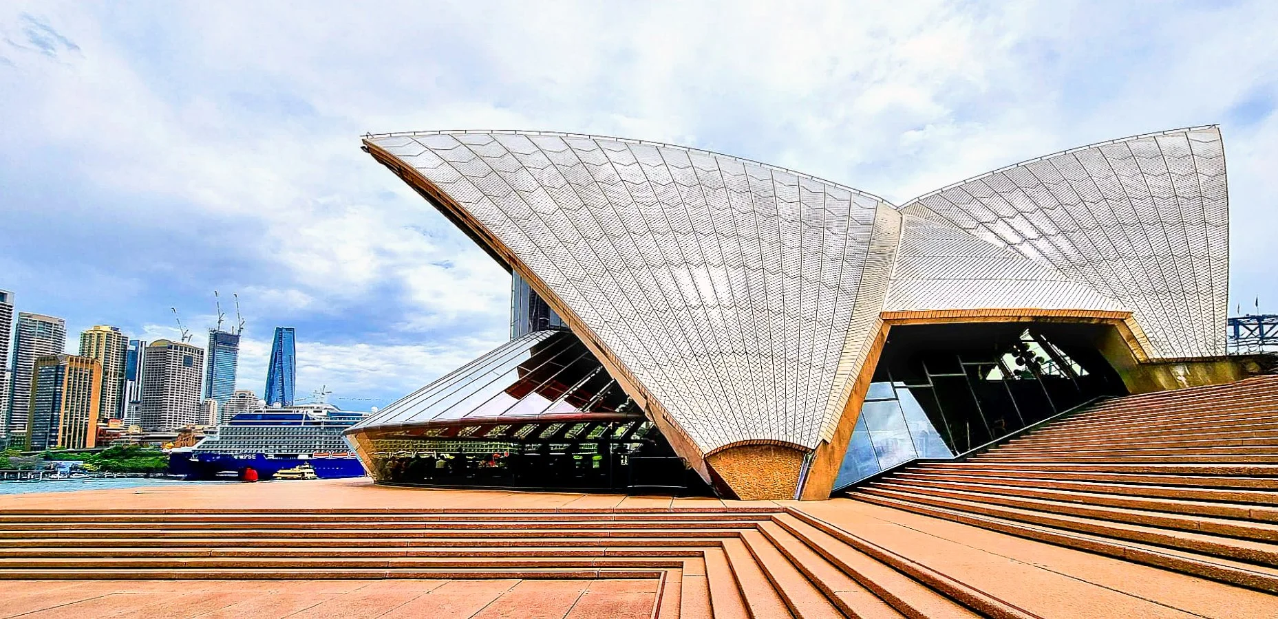 Sydney Opera House with steps leading up to it, city skyline with tall buildings and cruise ships in the harbor under cloudy sky.