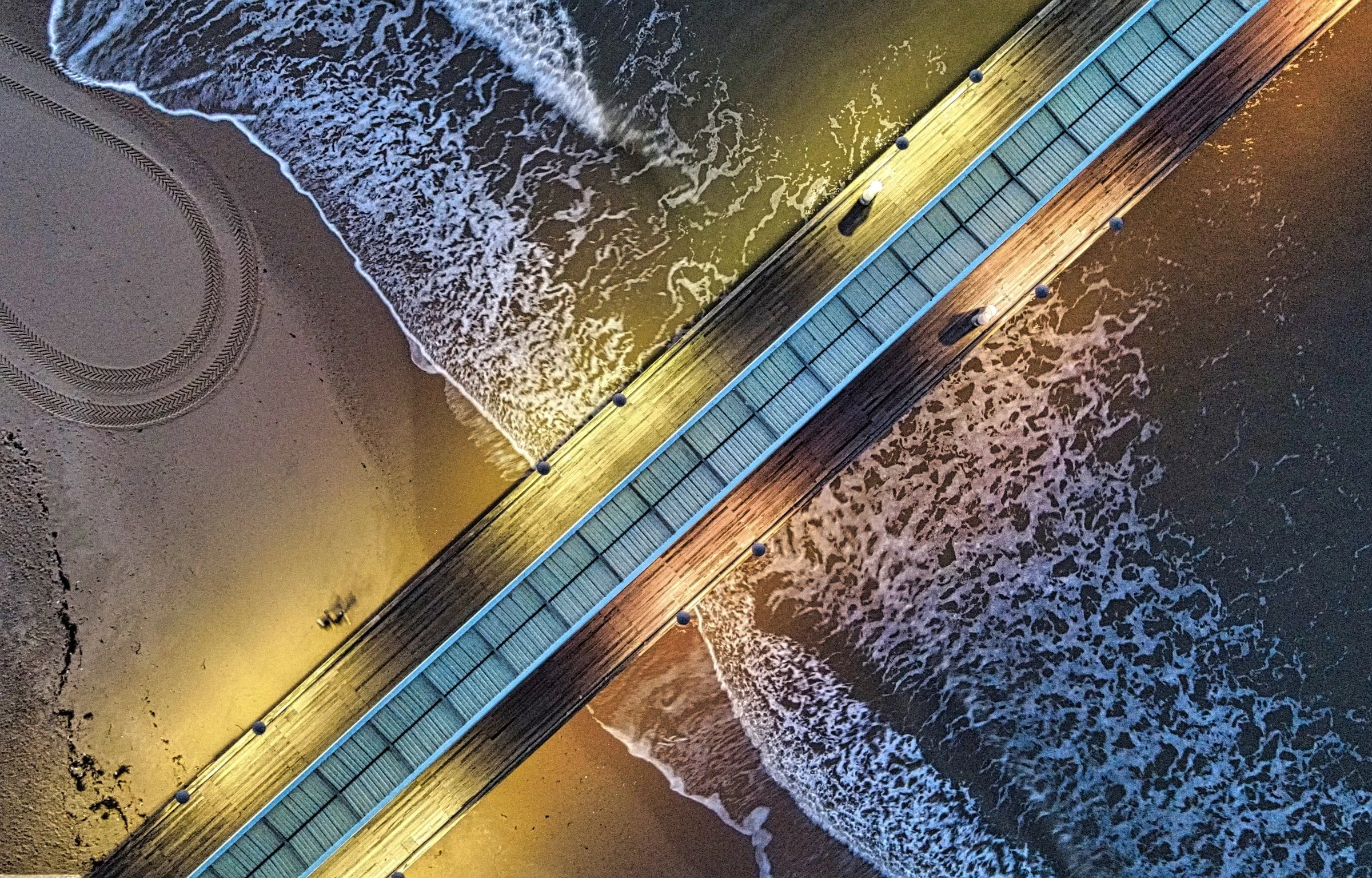Bird's eye view of a pier extending from the sandy beach into the ocean, with waves crashing against the shore at night illuminated by artificial lights.