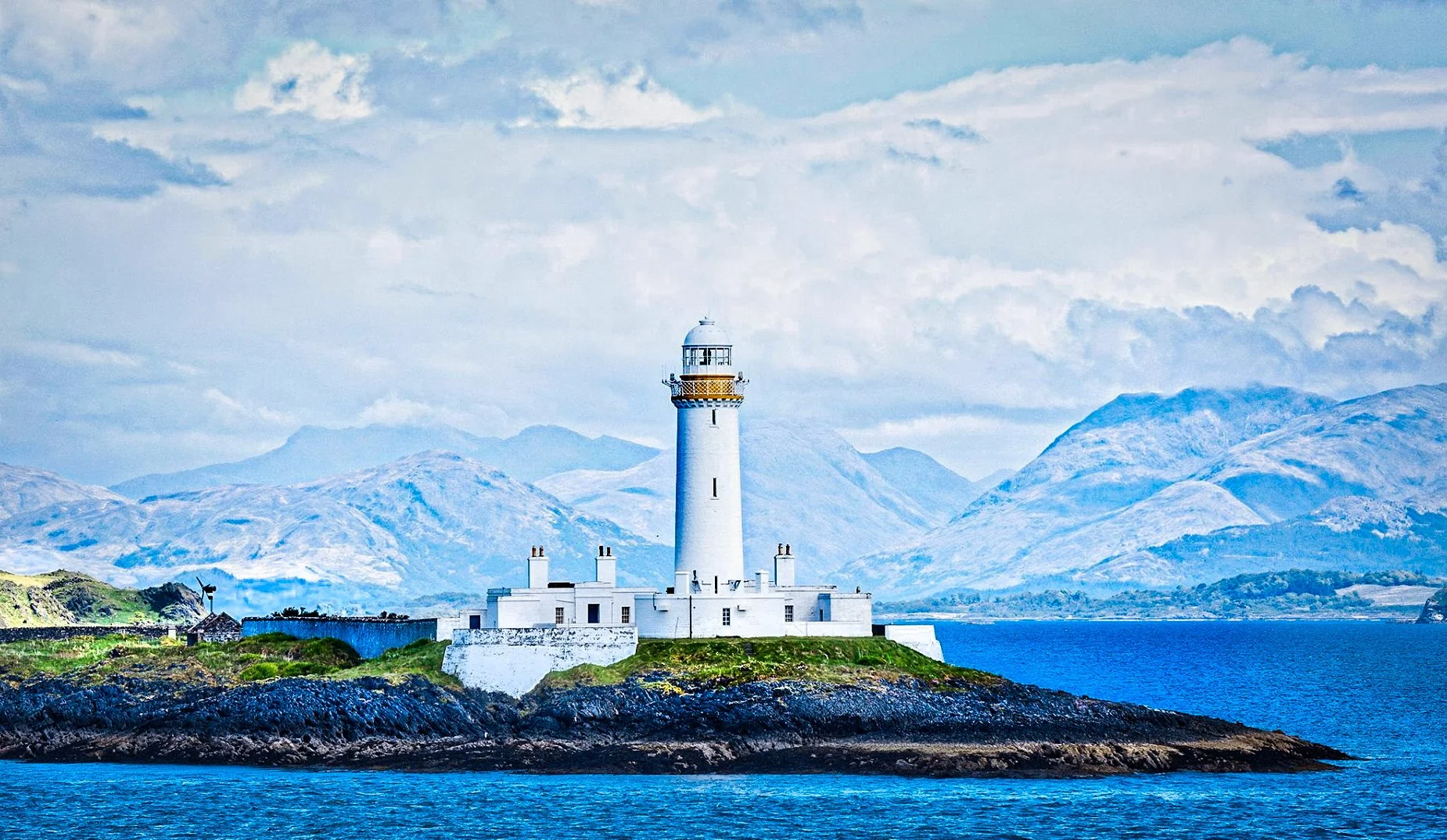 A white lighthouse on a small island with a castle-like base, surrounded by water, with mountains in the background under a partly cloudy sky.