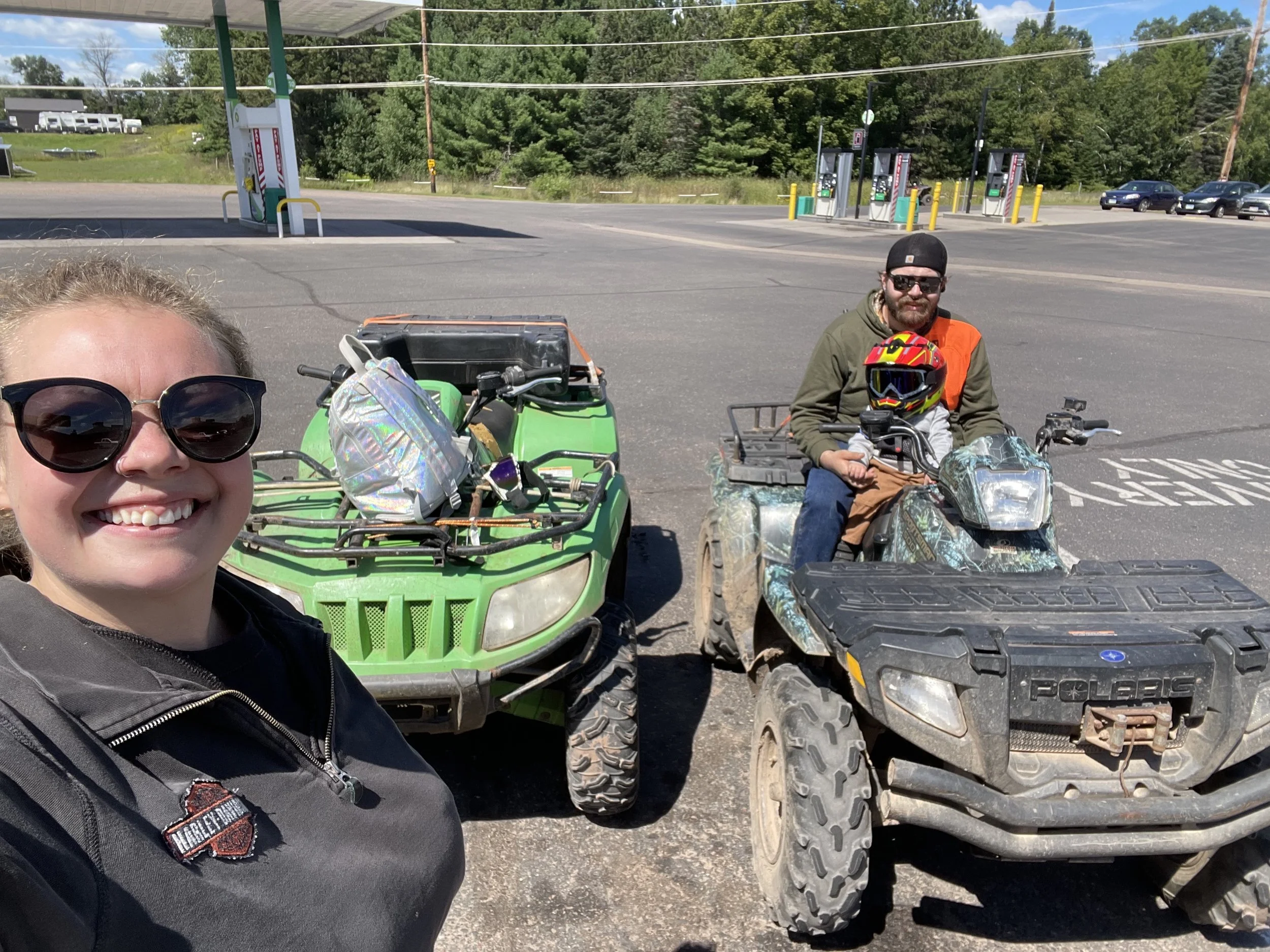 A smiling family, including a woman with sunglasses, takes a selfie in front of two all-terrain vehicles, with a man and a child on one of the vehicles, at a gas station on a sunny day with trees in the background.
