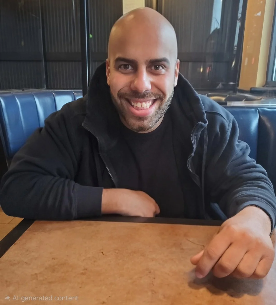 A smiling man with a shaved head and beard sitting at a table in a restaurant or cafe, wearing a black jacket.