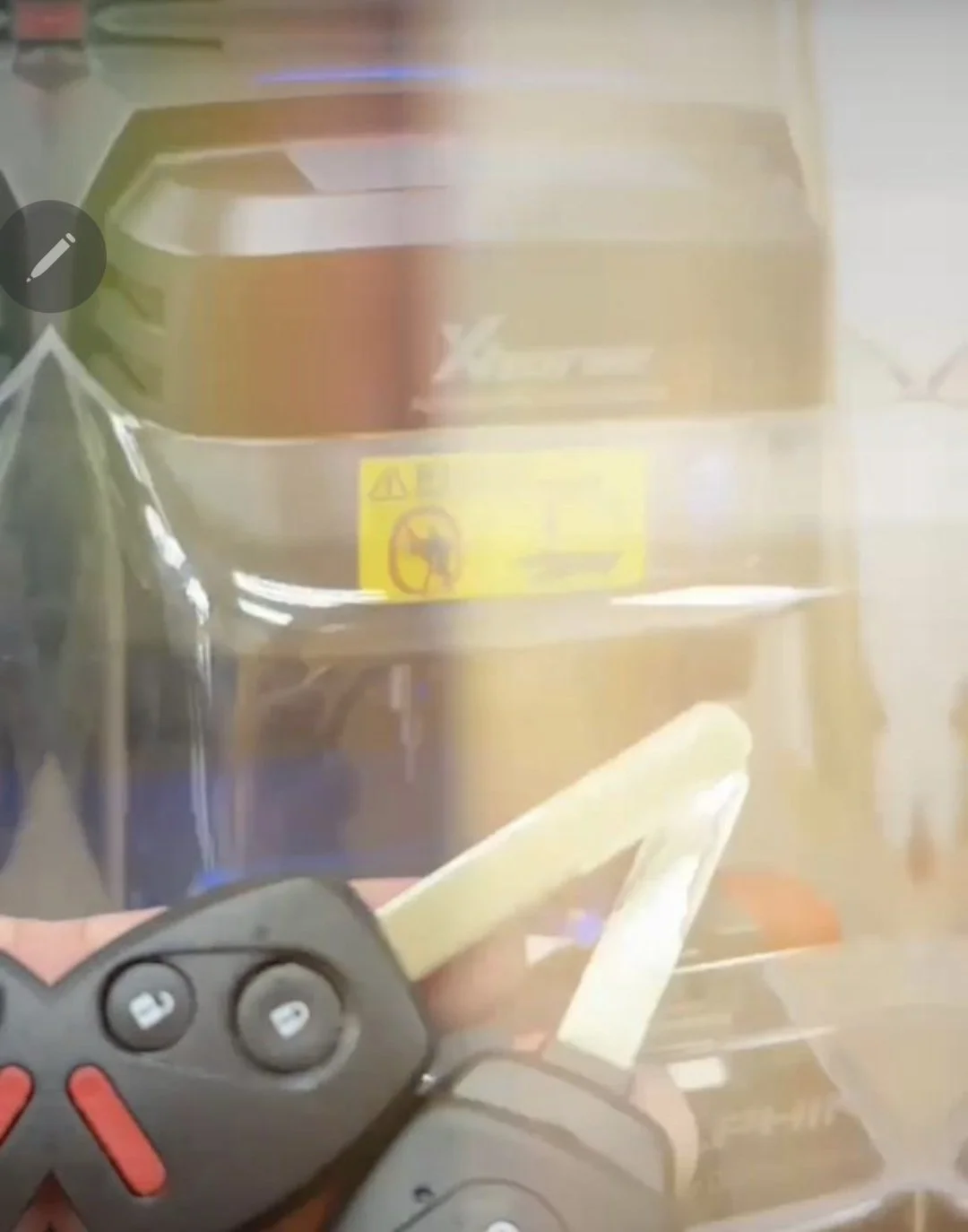 Close-up of a car key fob with buttons for lock, unlock, and trunk, held near a plastic container with a yellow warning label, in front of a stove with a vent hood.