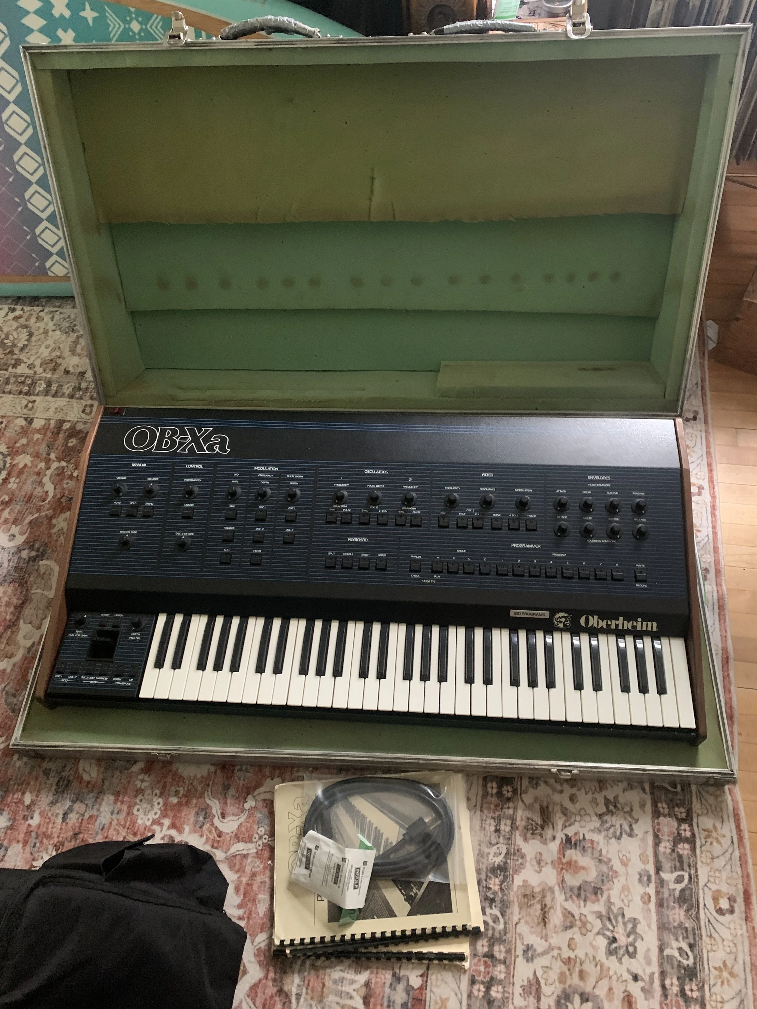 A vintage Oberheim OB-Xa synthesizer with a black control panel, white keys, and a green foam interior of its case. The case is open and placed on a patterned area rug, with a small manual and a cable in front of it, and a black bag partially visible in the lower left corner.