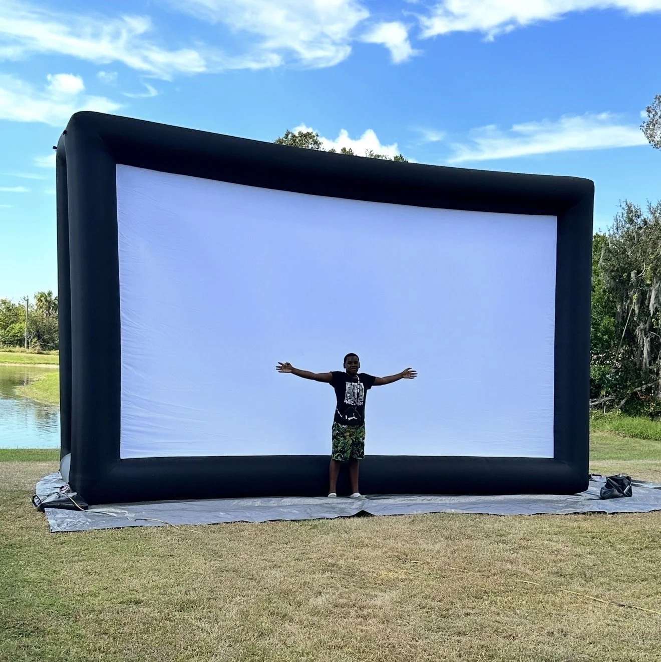 A person standing in front of a large inflatable movie screen outdoors on grass with arms outstretched.
