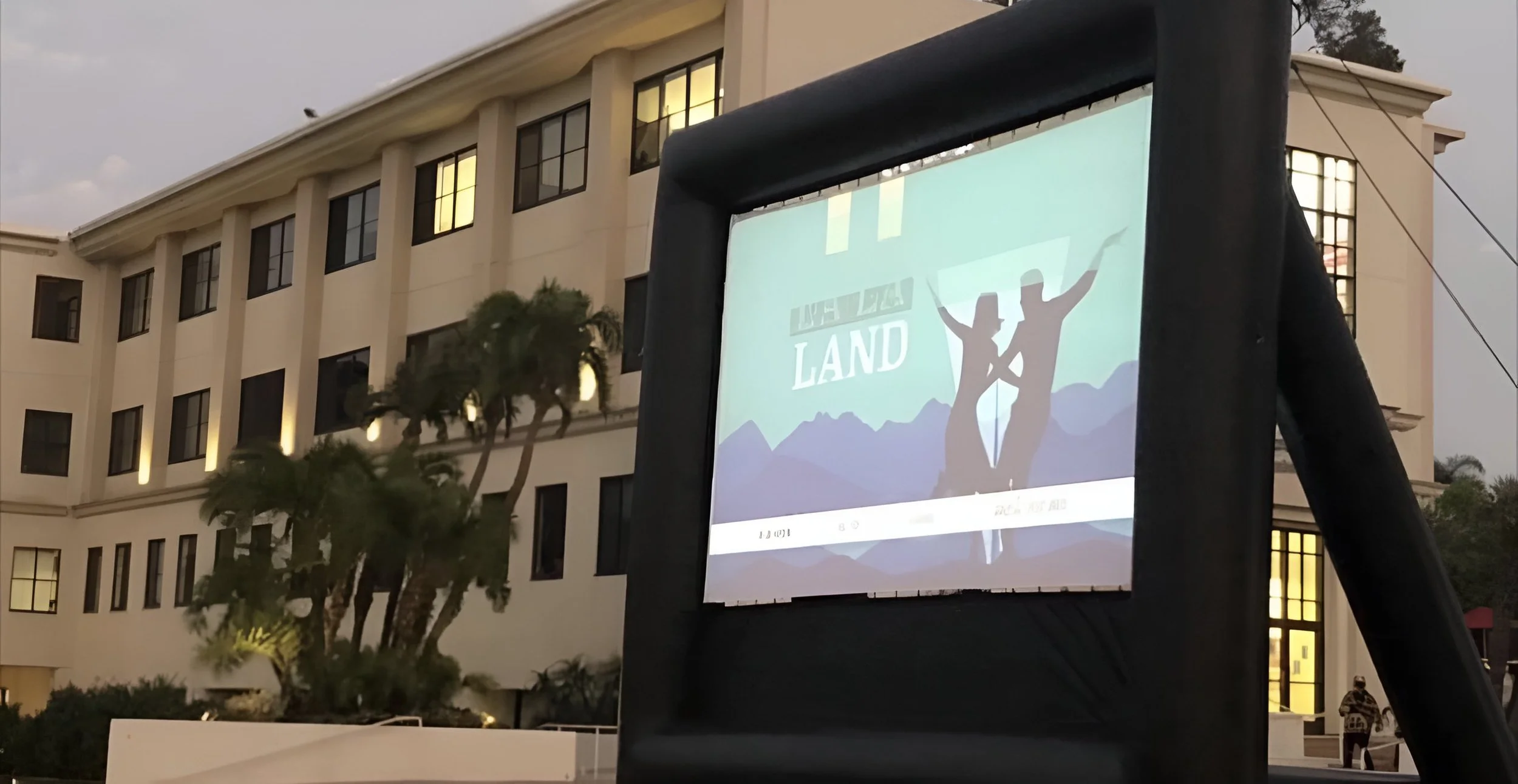 An outdoor movie screen displaying a promotional poster for the film 'Land,' with a silhouette of two people dancing against a mountain background, in front of a large building with lit windows at dusk.