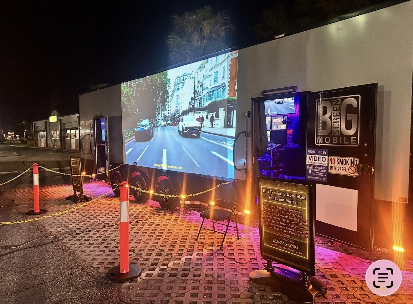 Mobile movie trailer with a large screen displaying a city street scene at night, surrounded by orange safety cones and chain barriers, with control equipment nearby and a sign advertising event rentals.