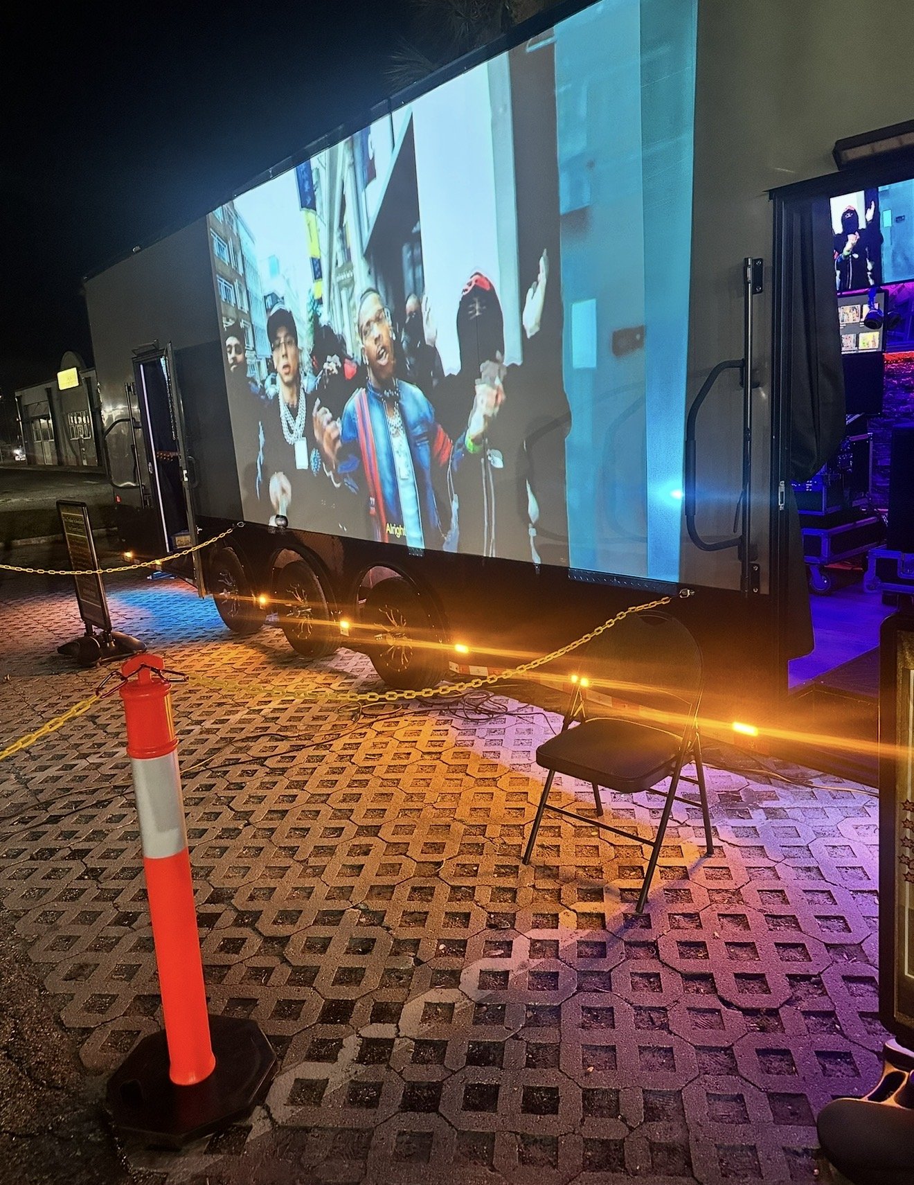 Large outdoor screen displaying a group of people walking in an urban area, with one person in front wearing a colorful jacket, holding a phone, and speaking, during nighttime. The screen is set on a street with a chain barrier, an orange and white traffic cone, a black chair, and illuminated pavement.