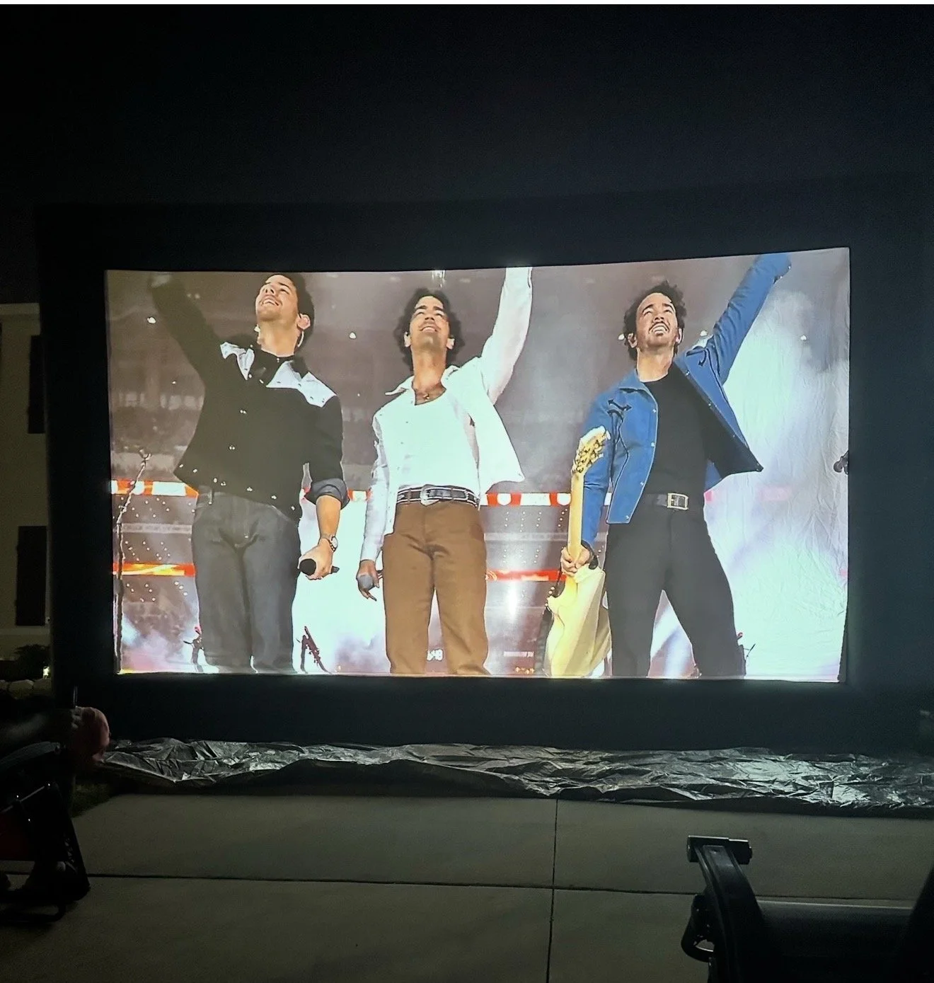 A large screen displaying a scene from a performance with three men on stage, holding microphones, smiling, and raising their arms.
