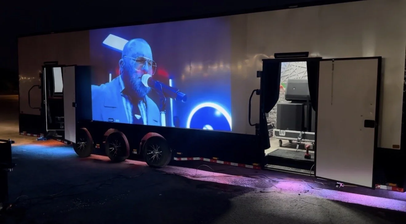 A large mobile LED screen on a trailer trailer displaying a man with a beard and glasses speaking into a microphone at night.