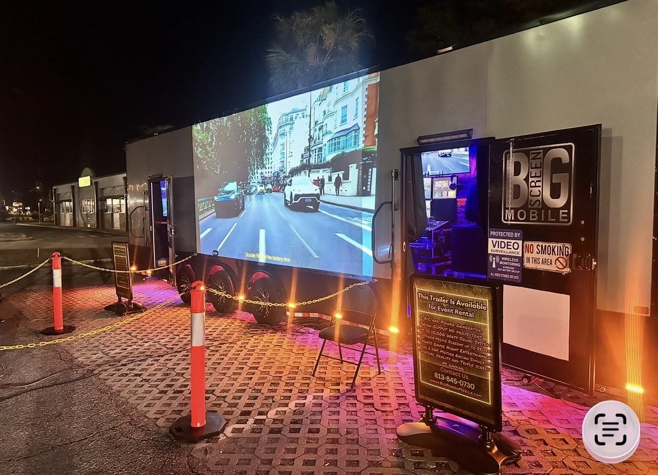 A portable screen showing a street view with cars and people at night, set up outdoors with safety posts and chain barriers, illuminated by pink and yellow lights.