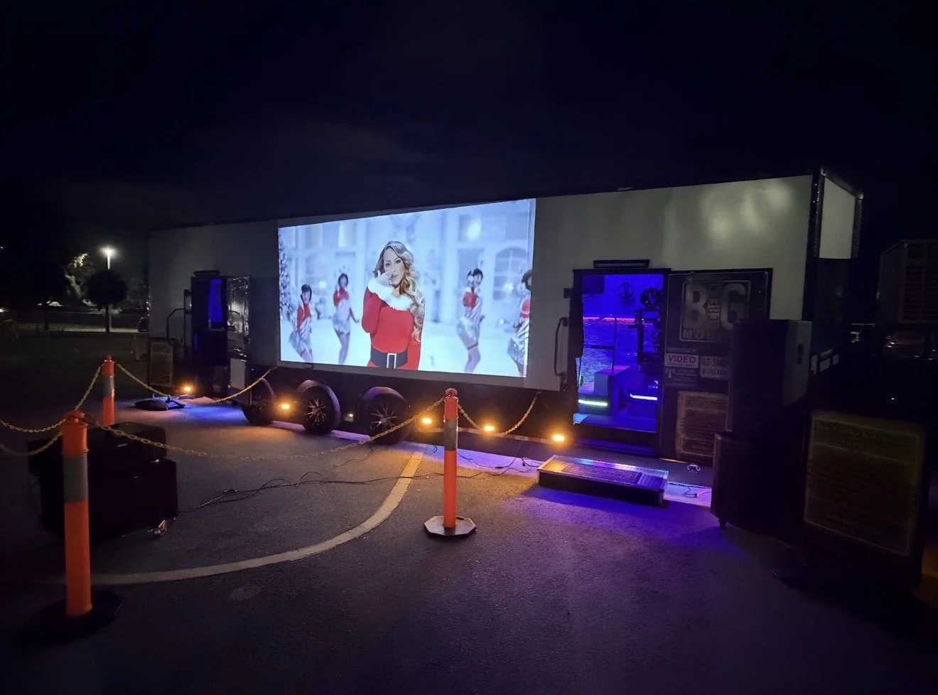 Outdoor mobile movie theater screen showing a woman in Christmas attire with dancers in the background, set up in a parking lot at night, surrounded by orange traffic cones and chains.