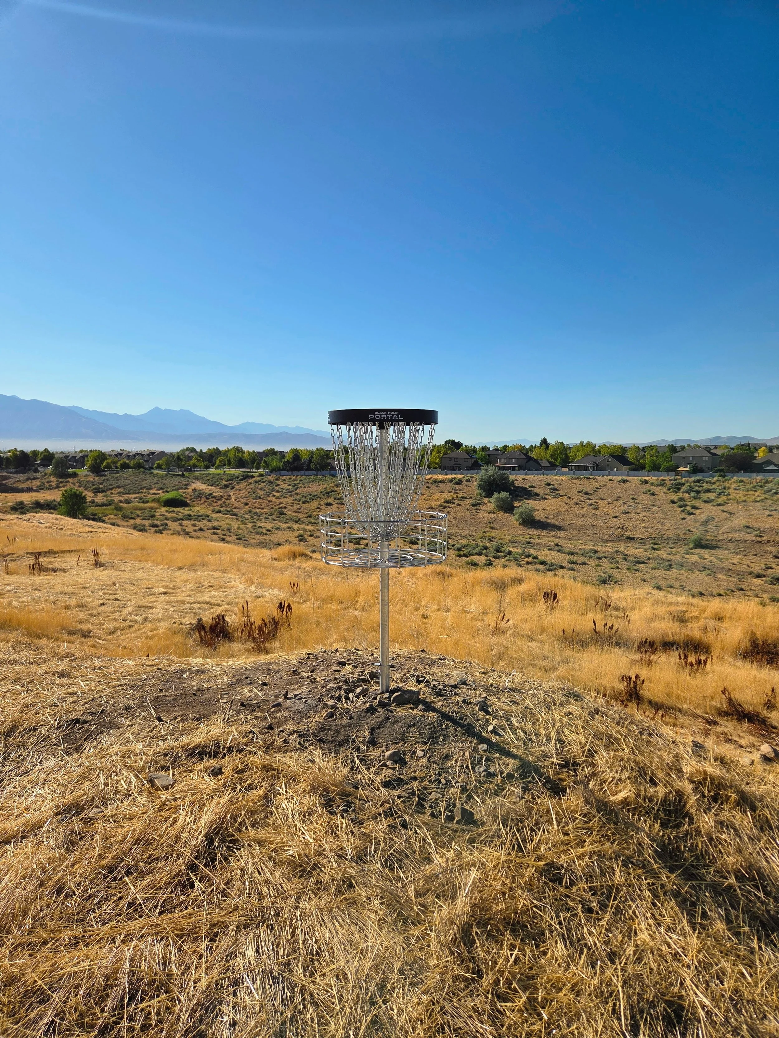 A disc golf basket in an open field with dry grass, houses in the background, and mountains under a clear blue sky.