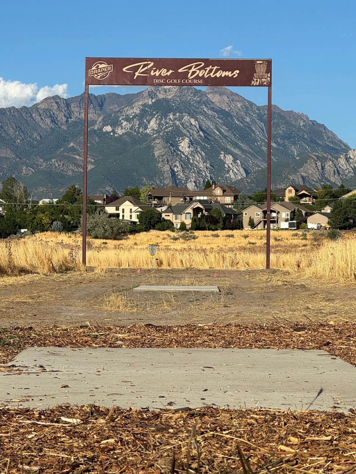 Sign for River Bottoms Disc Golf Course with a view of mountains in the background, houses, and a field of dry grass.