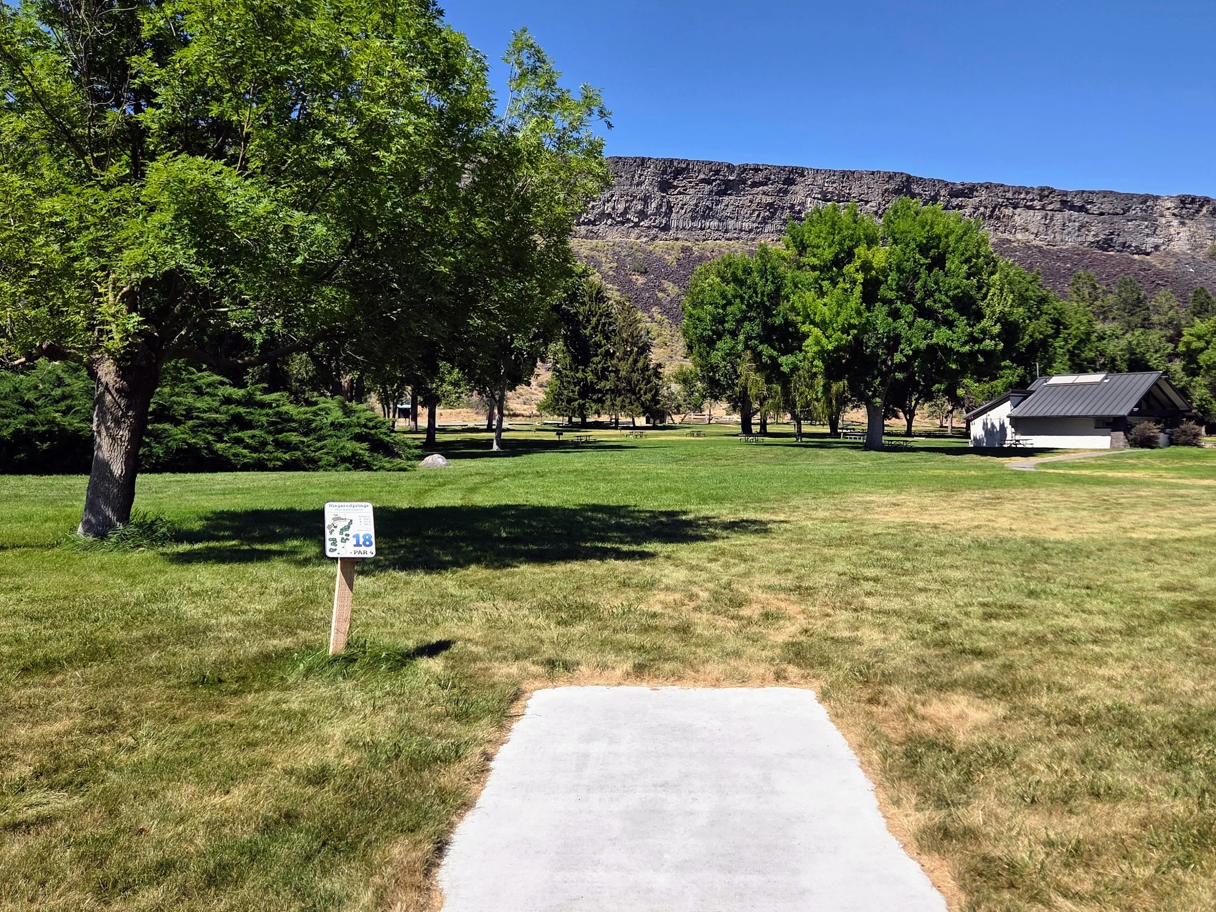 A park scene with green grass, trees, picnic tables, and a small building. A sign indicates it is Niagara Springs, 18th hole, par 4, part of a golf course. In the background, a rocky hillside is visible under a clear blue sky.