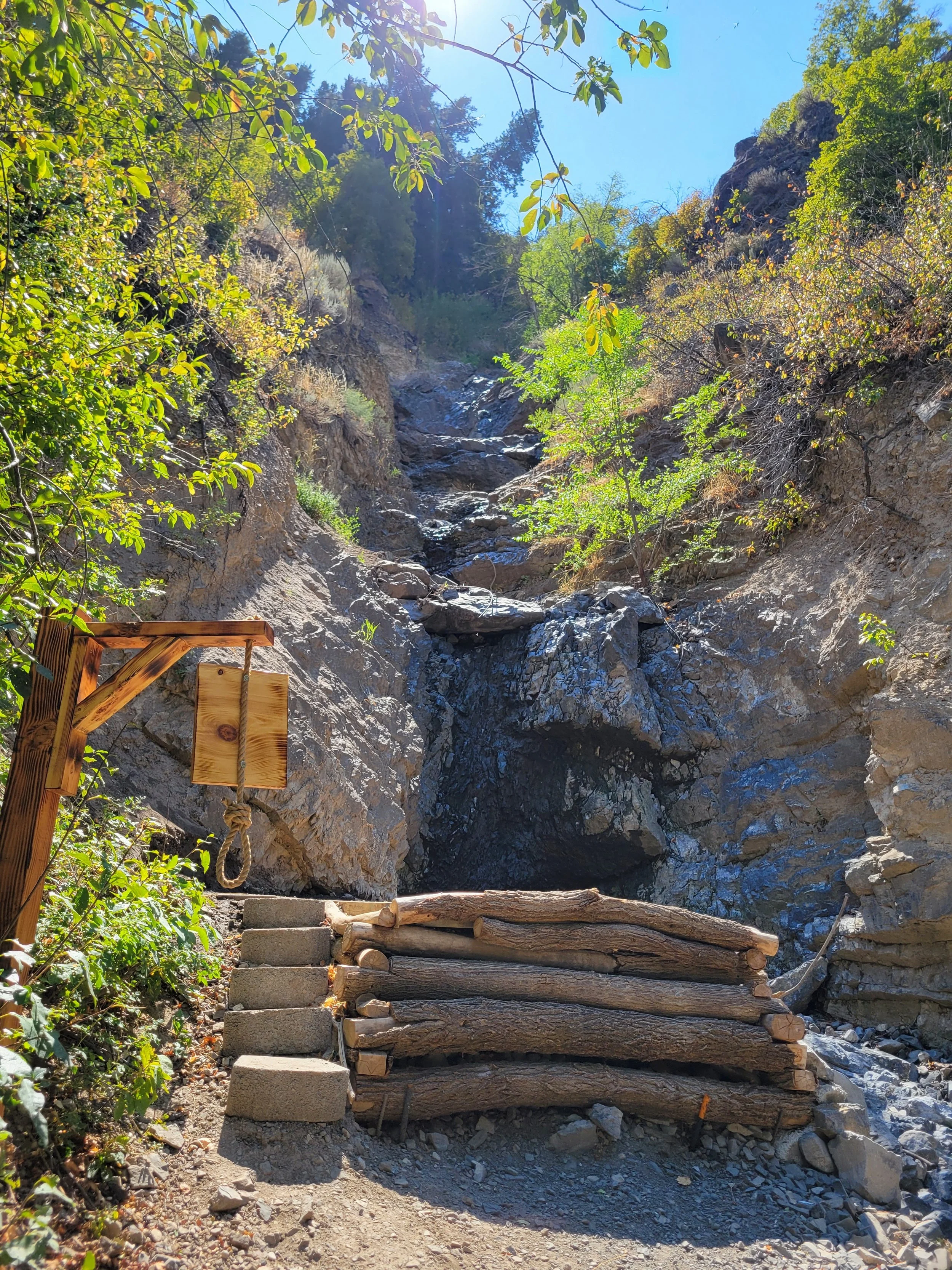 A rocky outdoor trail with a makeshift wooden staircase leading up a mountain or hillside. Bright daylight filters through green trees and bushes at the top of the scene, with a clear blue sky above.