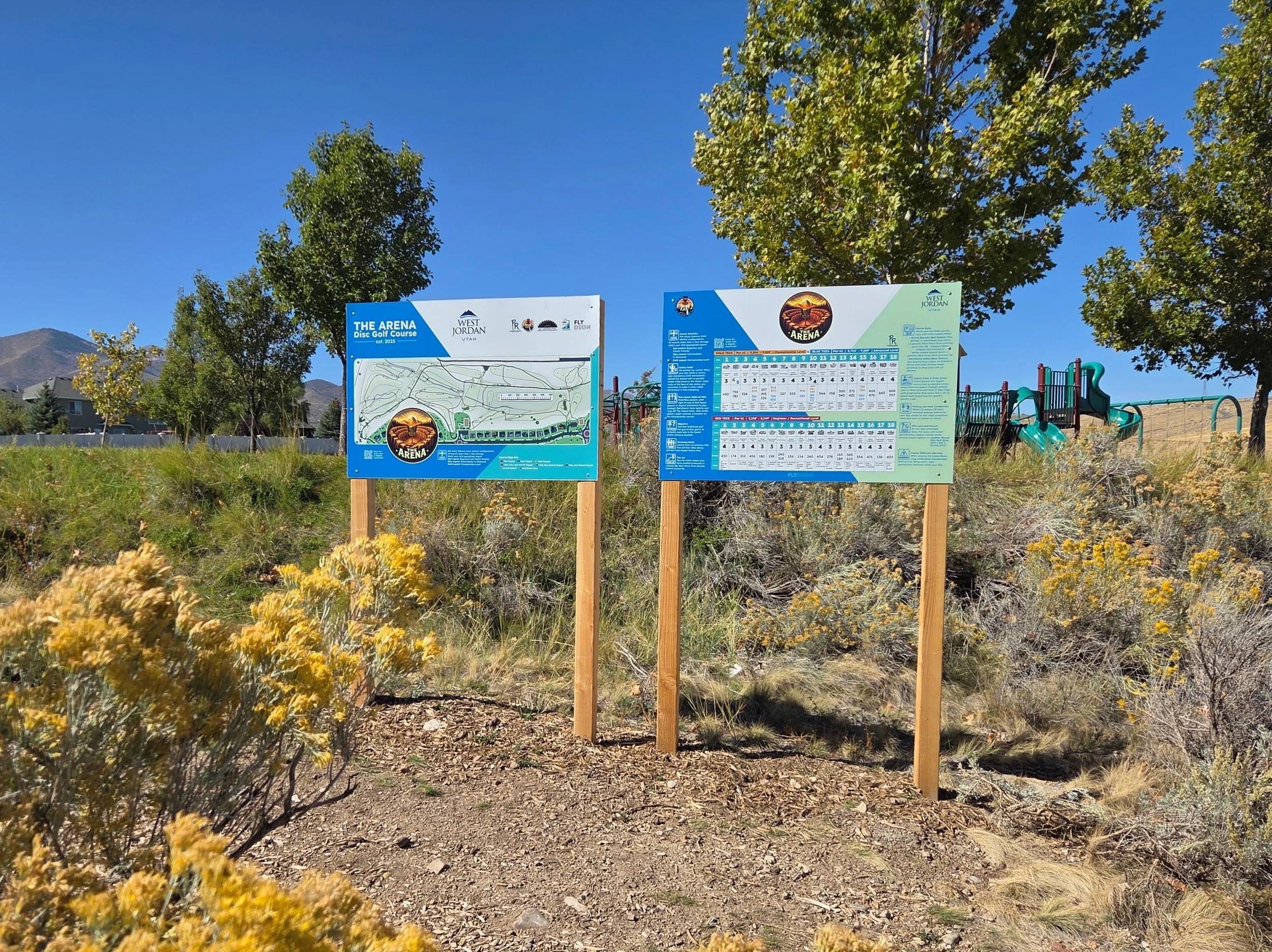 Signage boards providing information about The Arena disc golf course, located in West Jordan, Utah, with a playground and trees in the background, and mountains in the distance under a clear blue sky.