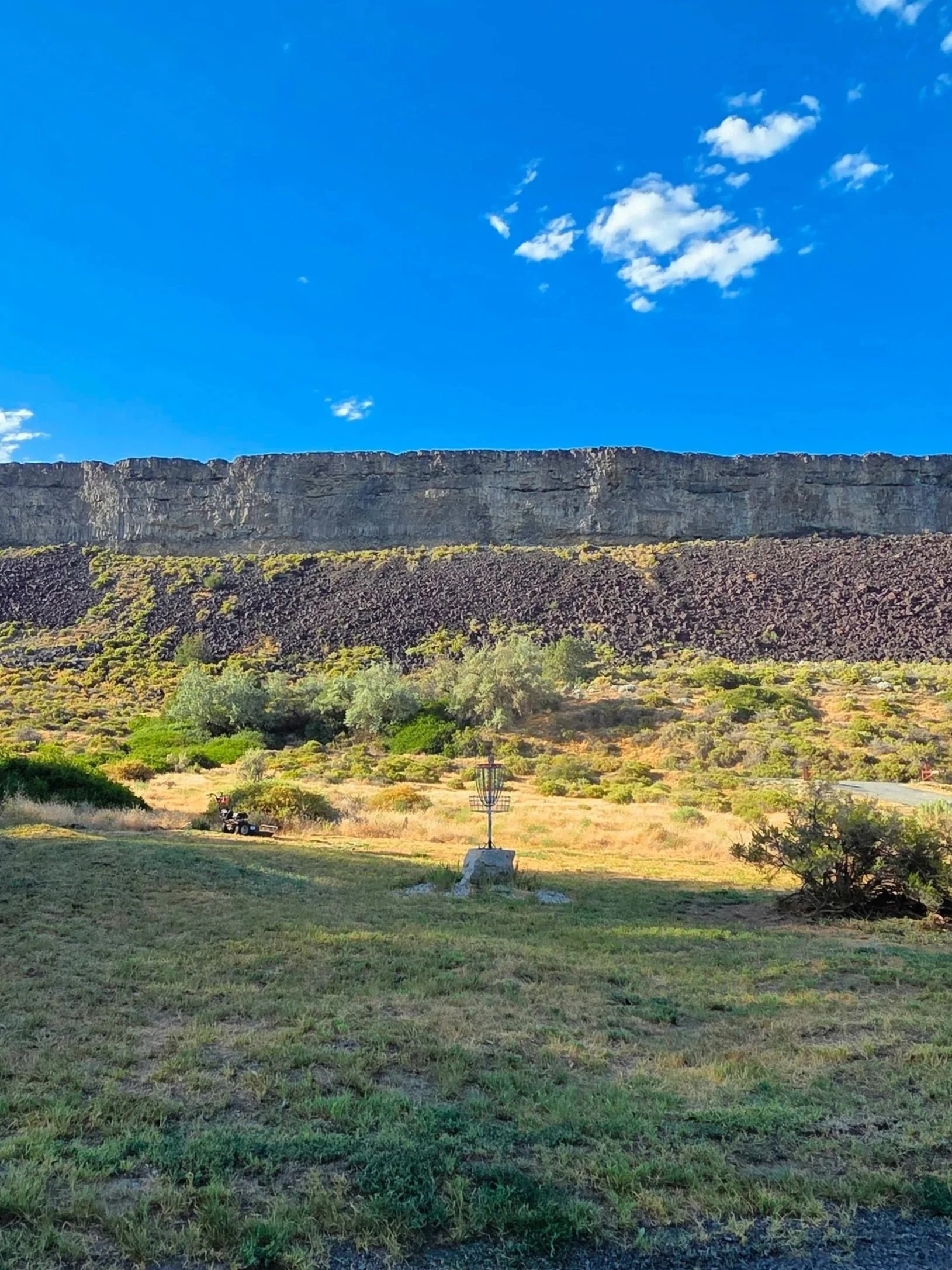 A landscape featuring a grassy foreground, a large rocky cliff in the middle, and a bright blue sky with some white clouds.