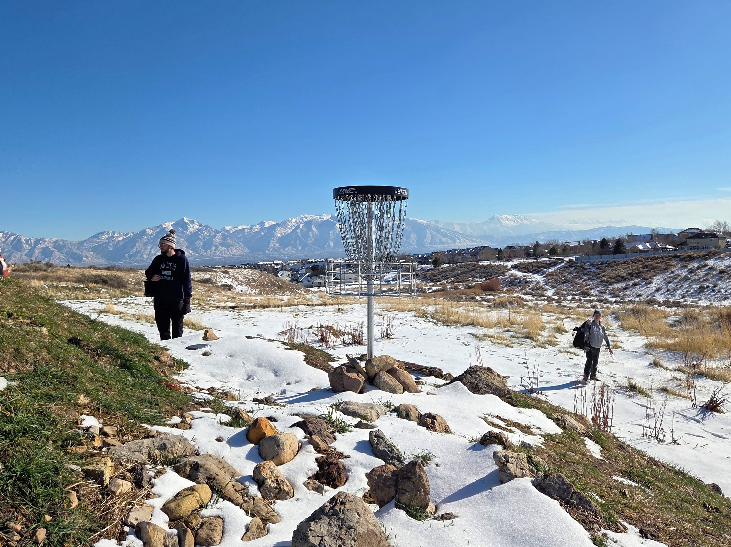 People walking on a snowy outdoor field with snow-covered mountains in the background, a disc golf basket in the foreground, and houses on the hillside.