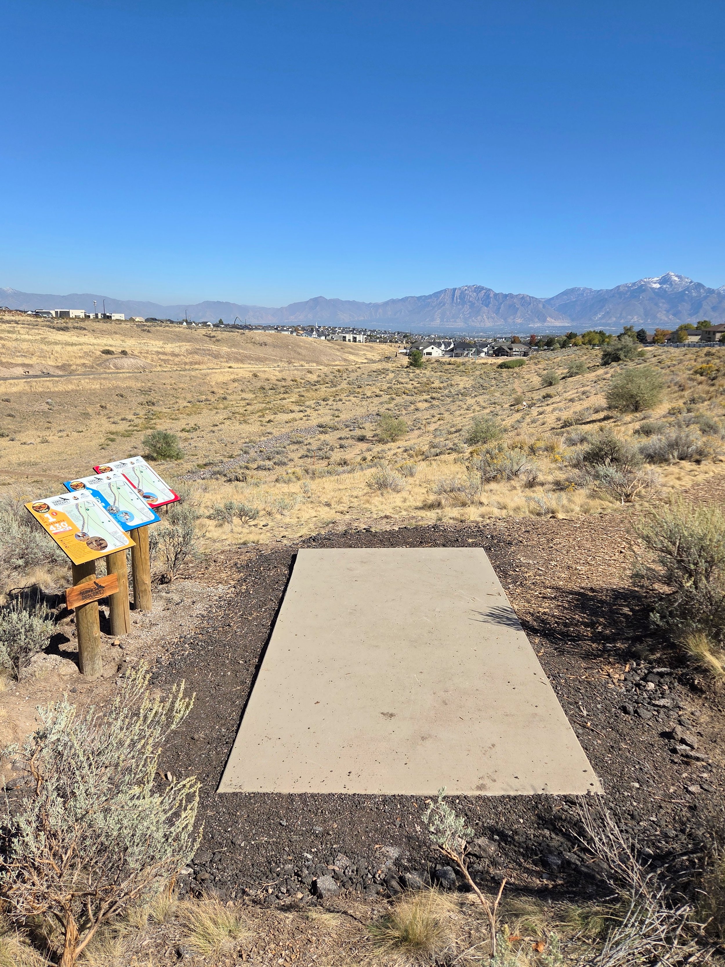 A concrete disc golf tee pad in a dry, grassy landscape with shrubs and mountains in the background under a clear blue sky, and a sign with instructions to the left.