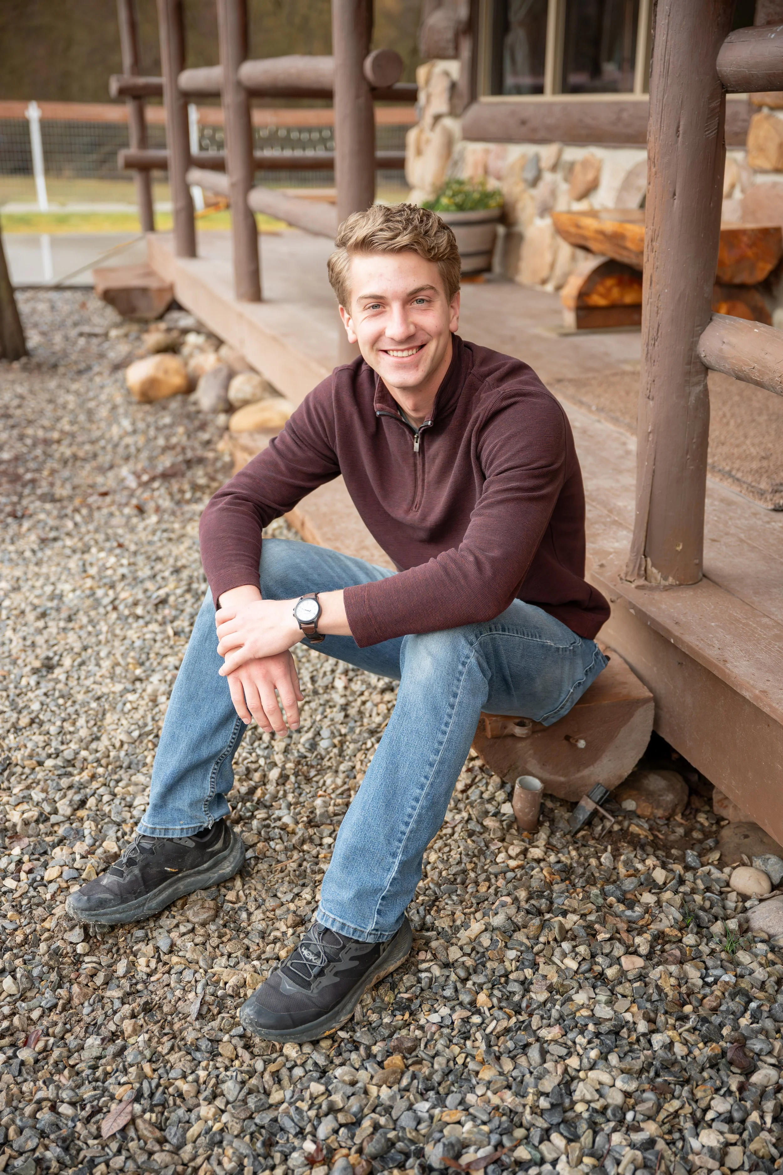 Young man with light brown hair, wearing a dark brown zip-up sweater, light blue jeans, black sneakers, and a watch, sitting on a wooden porch with a stone foundation, surrounded by gravel and small rocks, smiling at the camera.