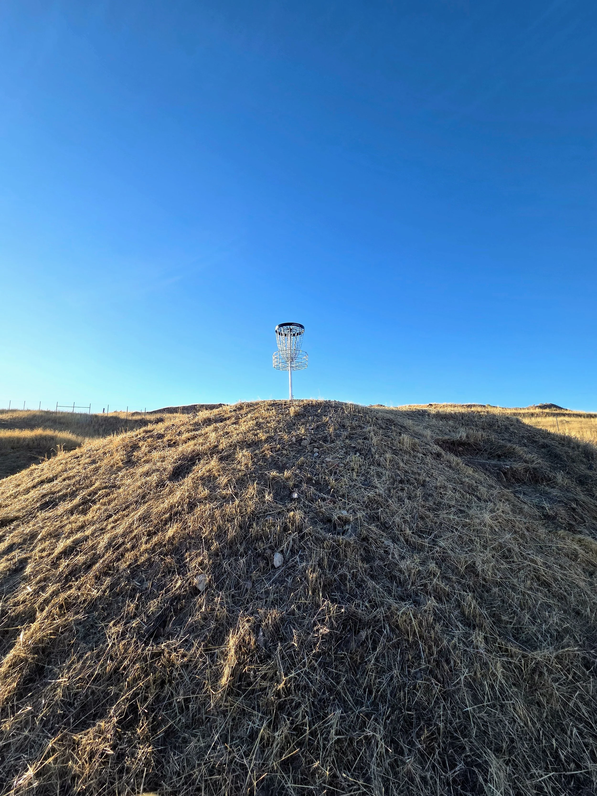 A disc golf basket atop a grassy hill under a clear blue sky.