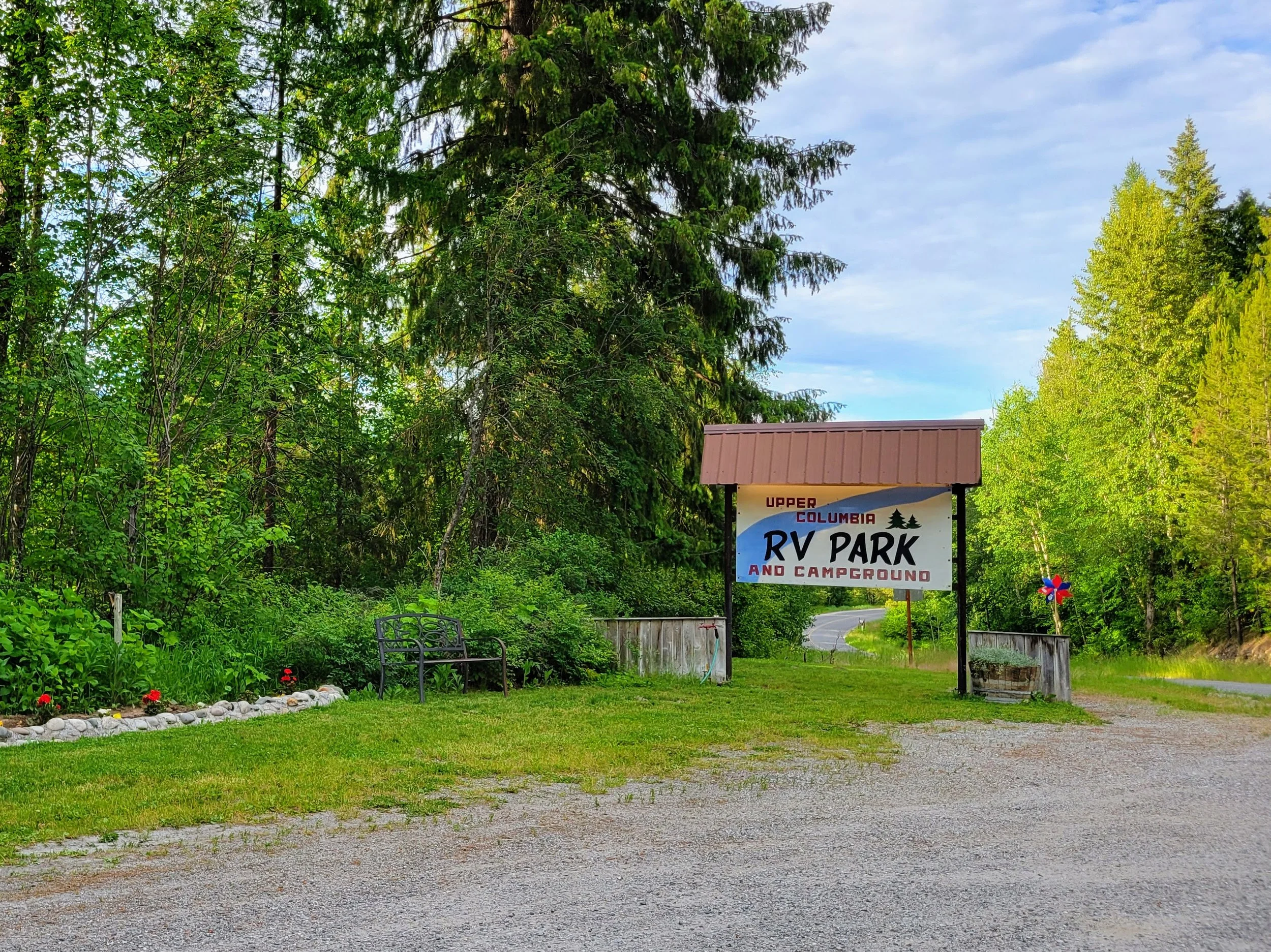 Entrance to Upper Columbia RV Park and Campground with sign, surrounded by green trees, a garden with flowers, and a gravel pathway.