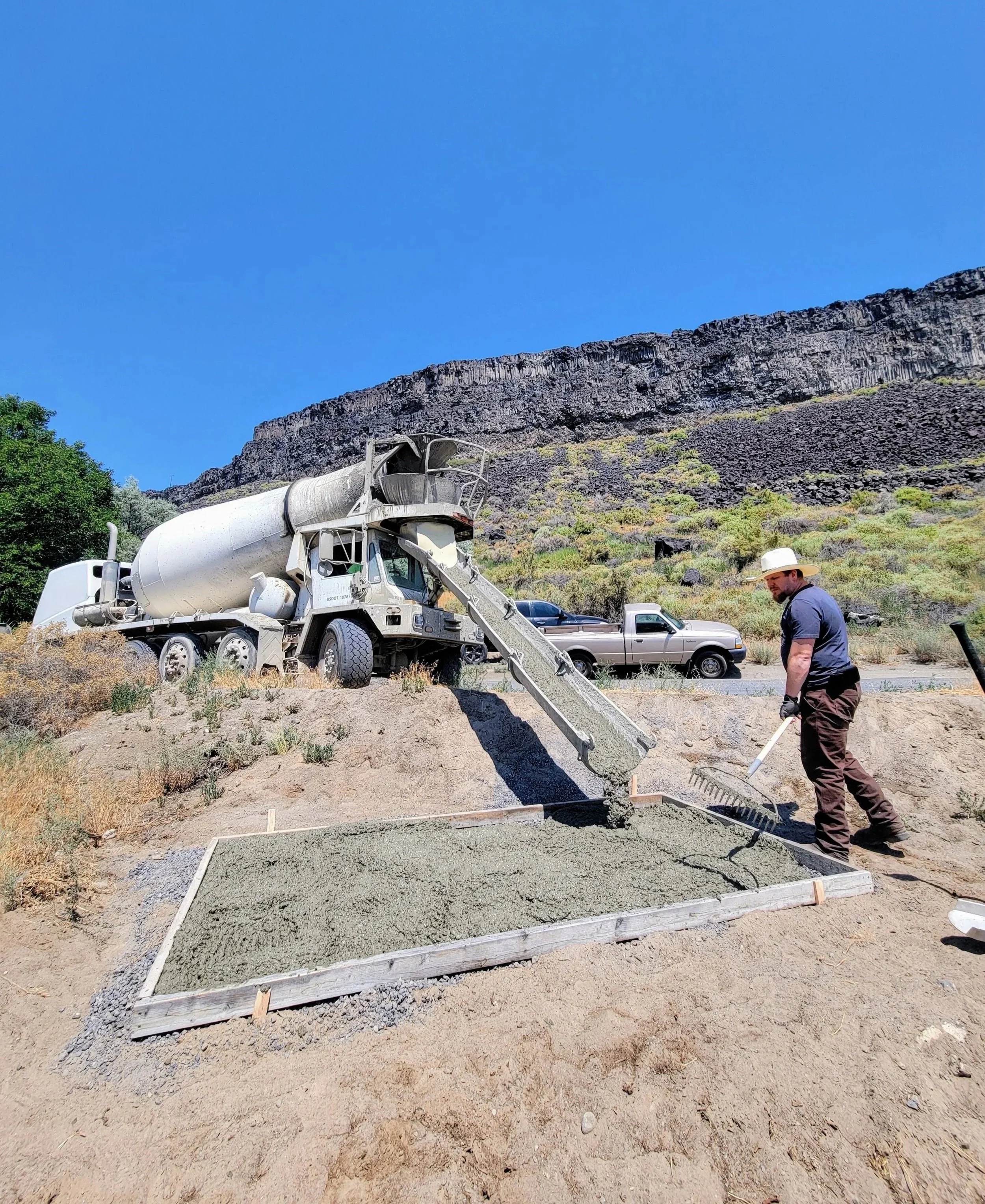 A construction site with a concrete mixing truck pouring concrete into a rectangular frame for a foundation, with a man wearing a hat and gloves smoothing the newly poured concrete. There's a rocky hillside and blue sky in the background.