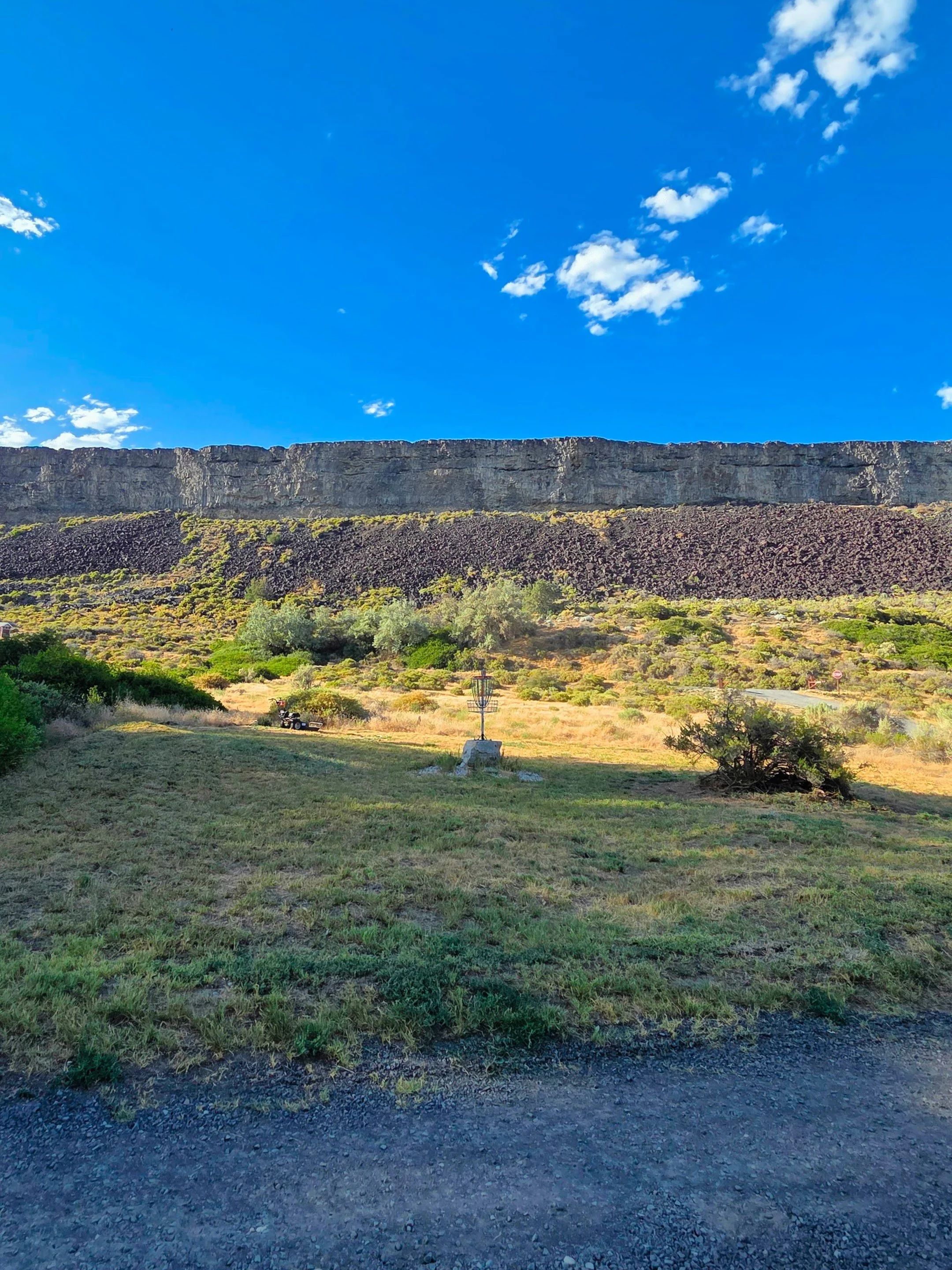 A landscape featuring a grassy foreground, a large rocky cliff in the middle, and a bright blue sky with some white clouds.