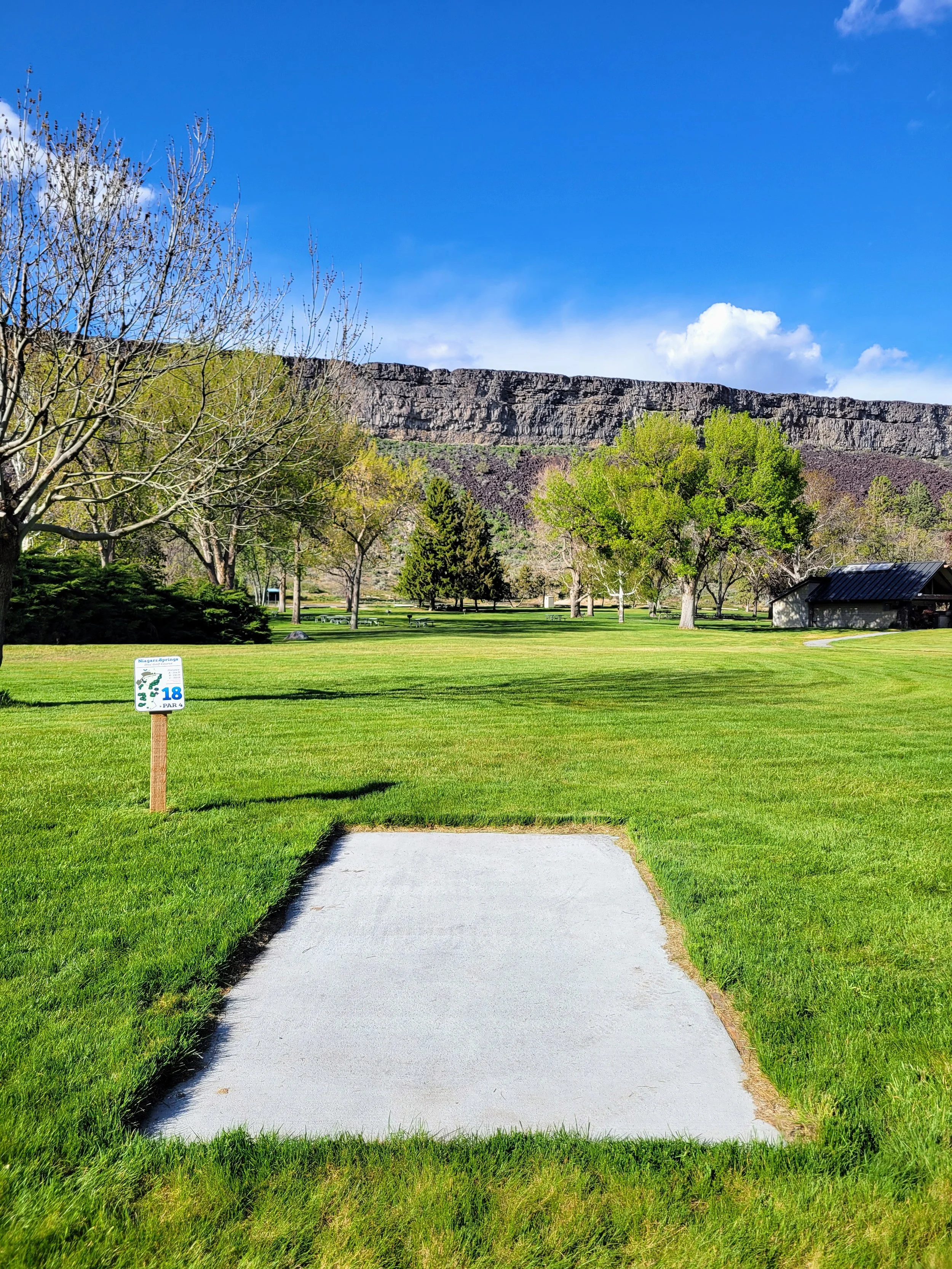 A well-maintained golf course with a tee box, lush green grass, trees in the background, and a sign indicating hole 18, with mountains and a blue sky with a few clouds in the distance.
