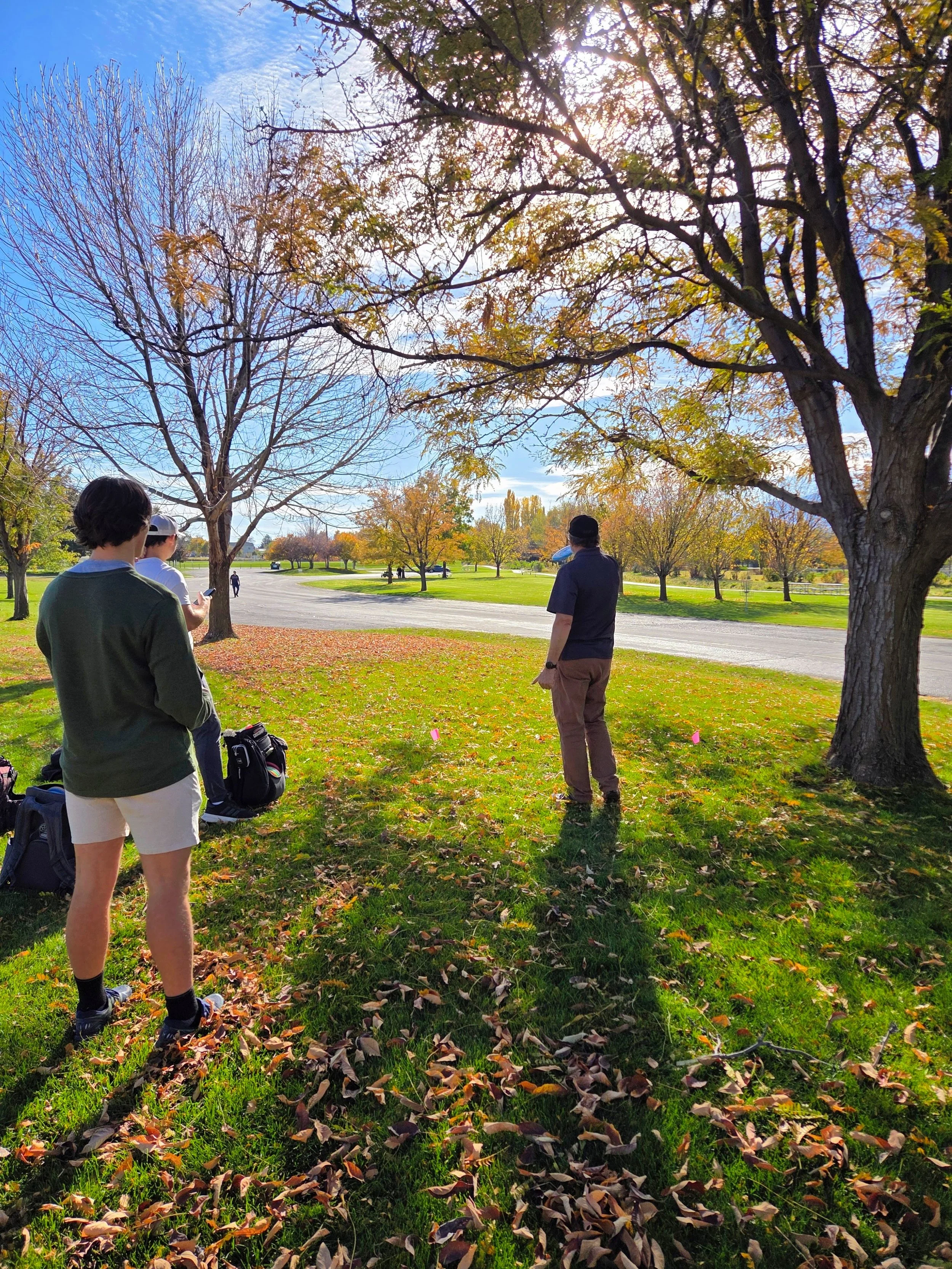 People standing outdoors in a park with autumn trees, some with colored leaves and some bare, on a sunny day with a bright sky.