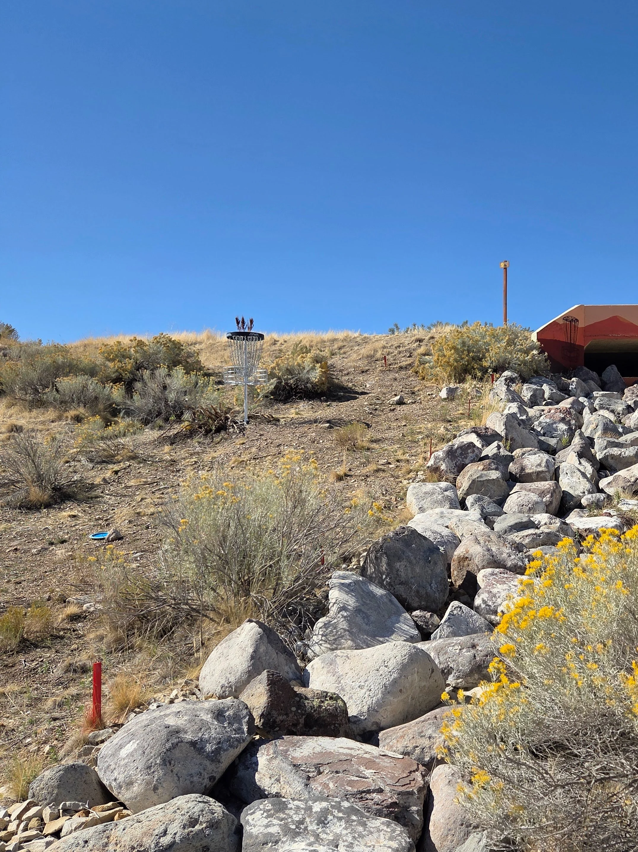 A disc golf basket on a rocky and desert-like landscape with yellow flowering plants, rocks, and a large red and white rectangular structure or shelter. The sky is clear and blue.