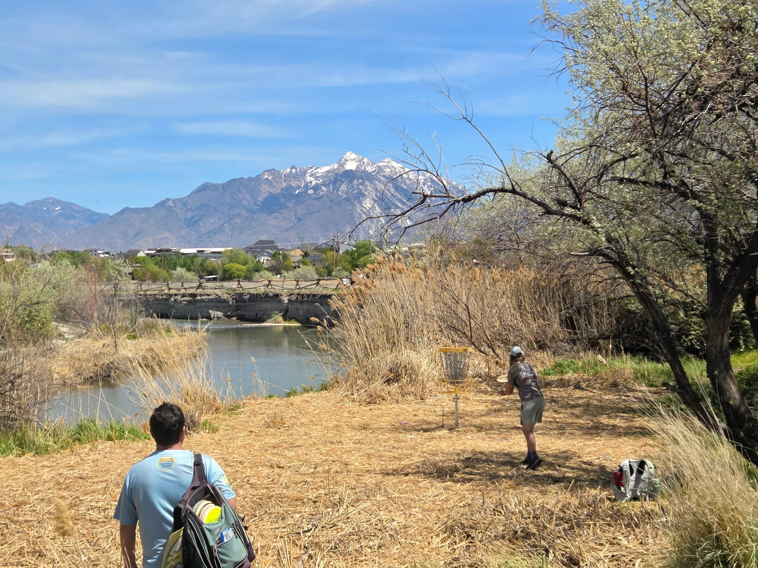 People observing and exploring nature by a river, with mountains in the background on a bright, clear day.