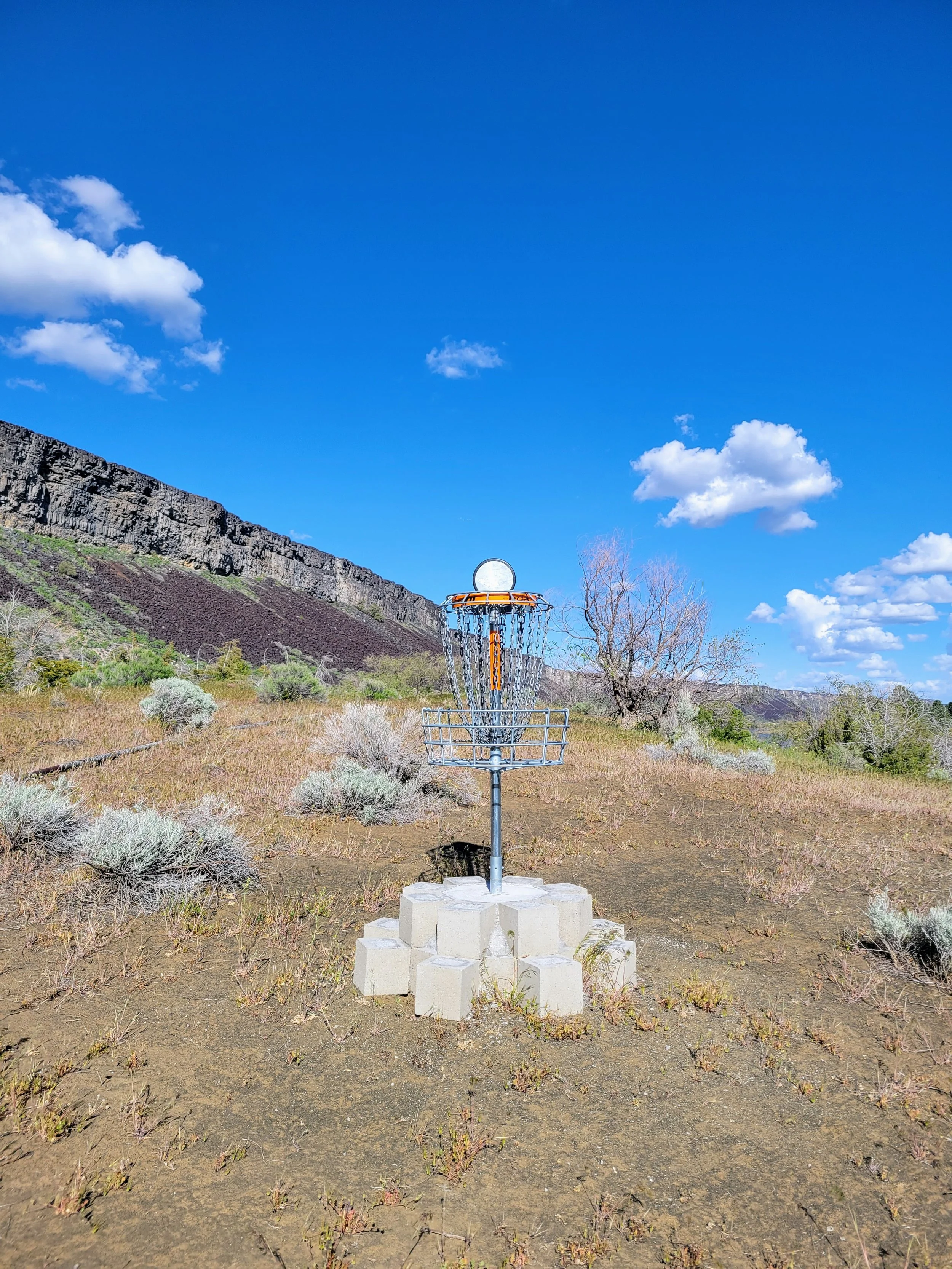 A disc golf basket with a metal chain assembly and a metal basket, set on a concrete base in a dry, grassy landscape with shrubs and a hill in the background, under a bright blue sky with scattered white clouds.