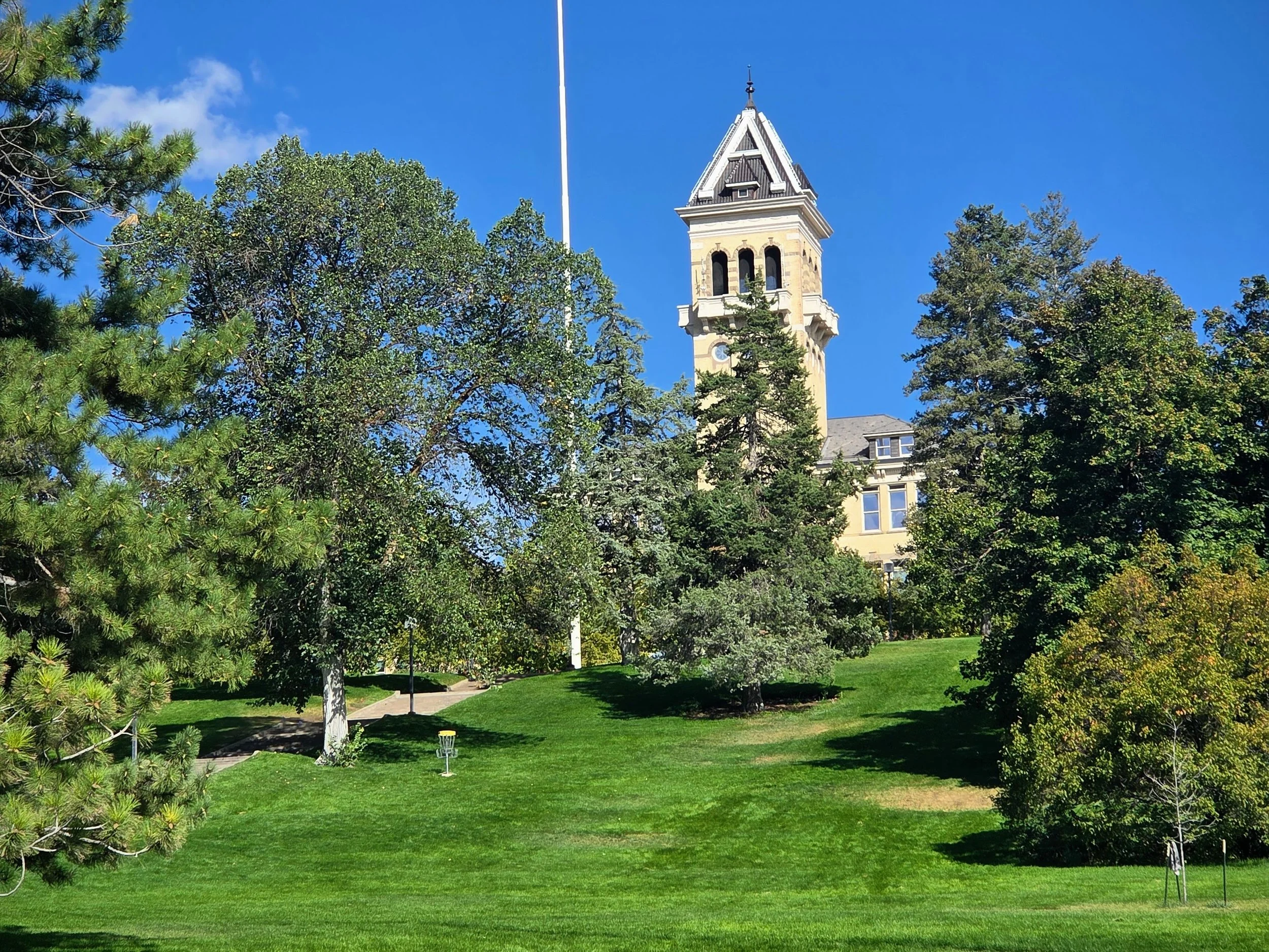 A park with green grass and various trees, a disc golf basket, and a tall clock tower with a pointed roof in the background under a clear blue sky. Utah State University disc golf course.
