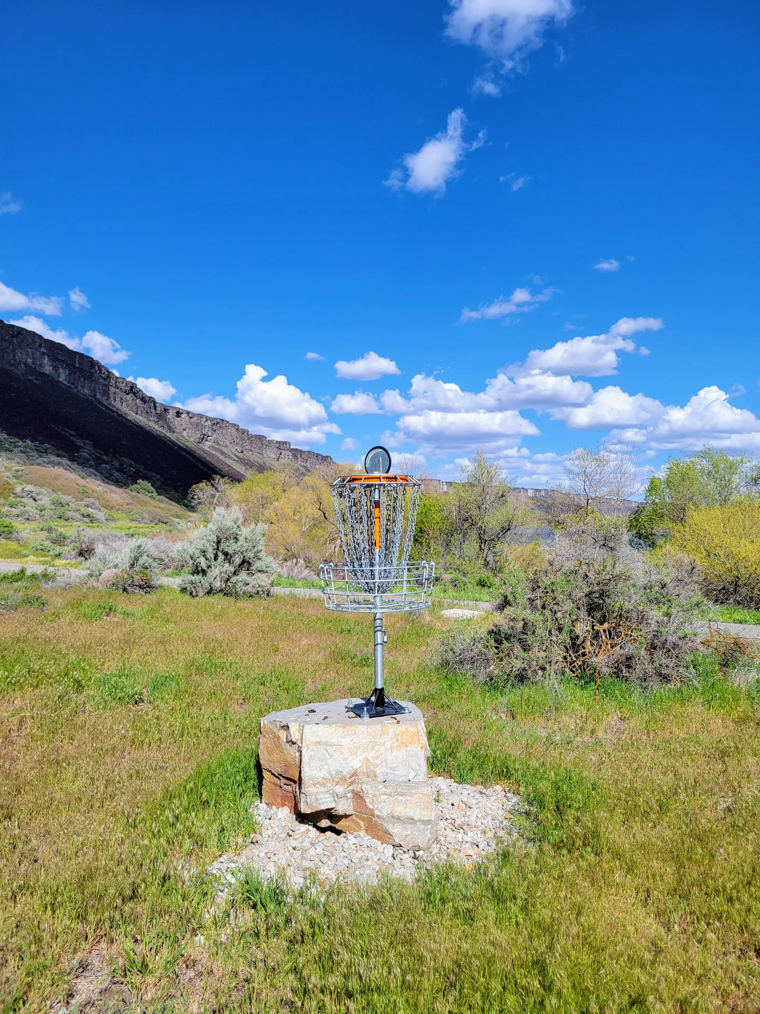 A disc golf basket situated outdoors on a stone base in a grassy field, with a mountainous landscape and blue sky with scattered clouds in the background.