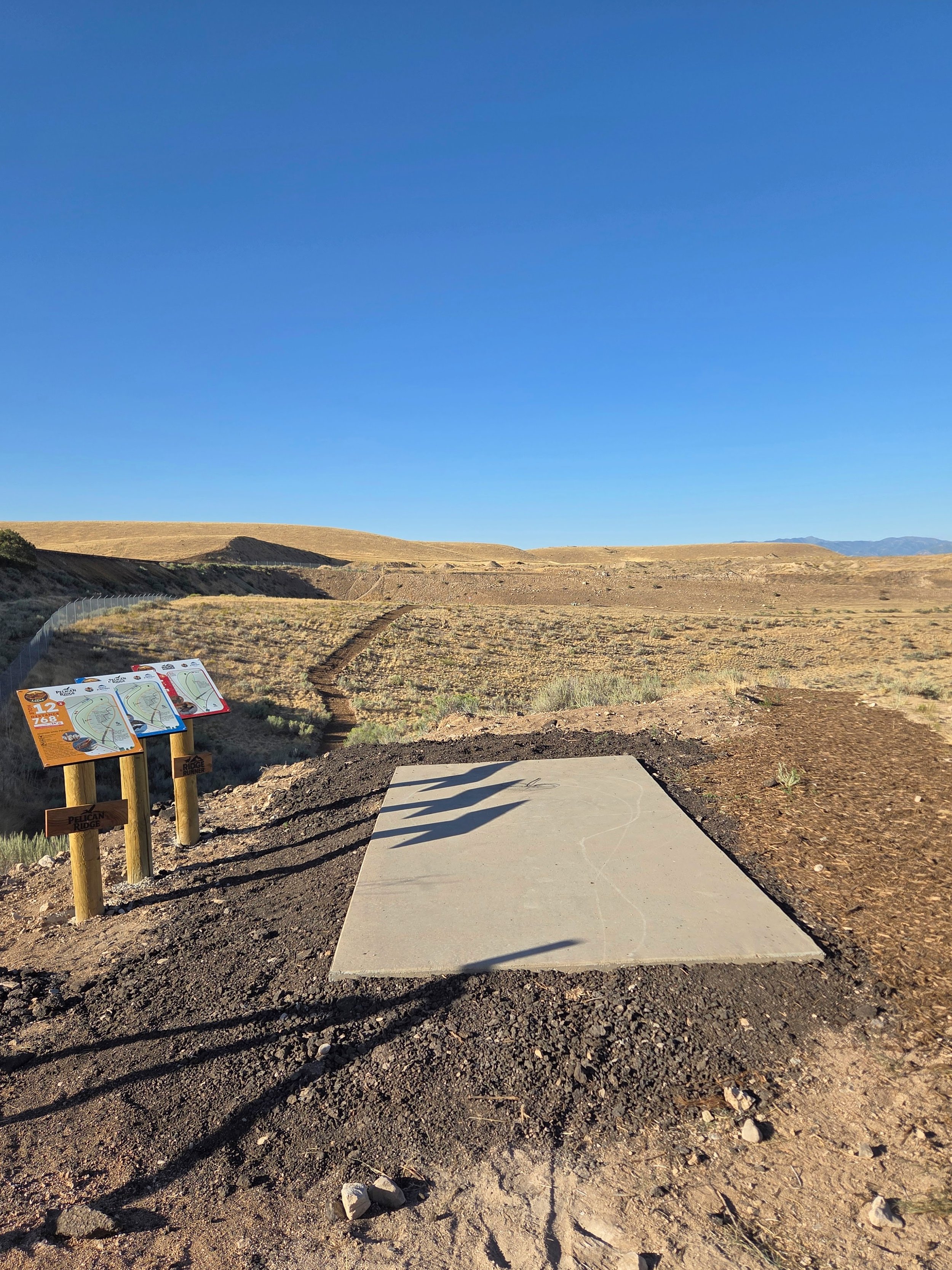 A flat, square concrete disc golf tee pad in a desert landscape with dry grass, dirt, and scattered rocks in the foreground. Nearby signs with map and course information are on wooden posts. Rolling hills and a clear blue sky are in the background.