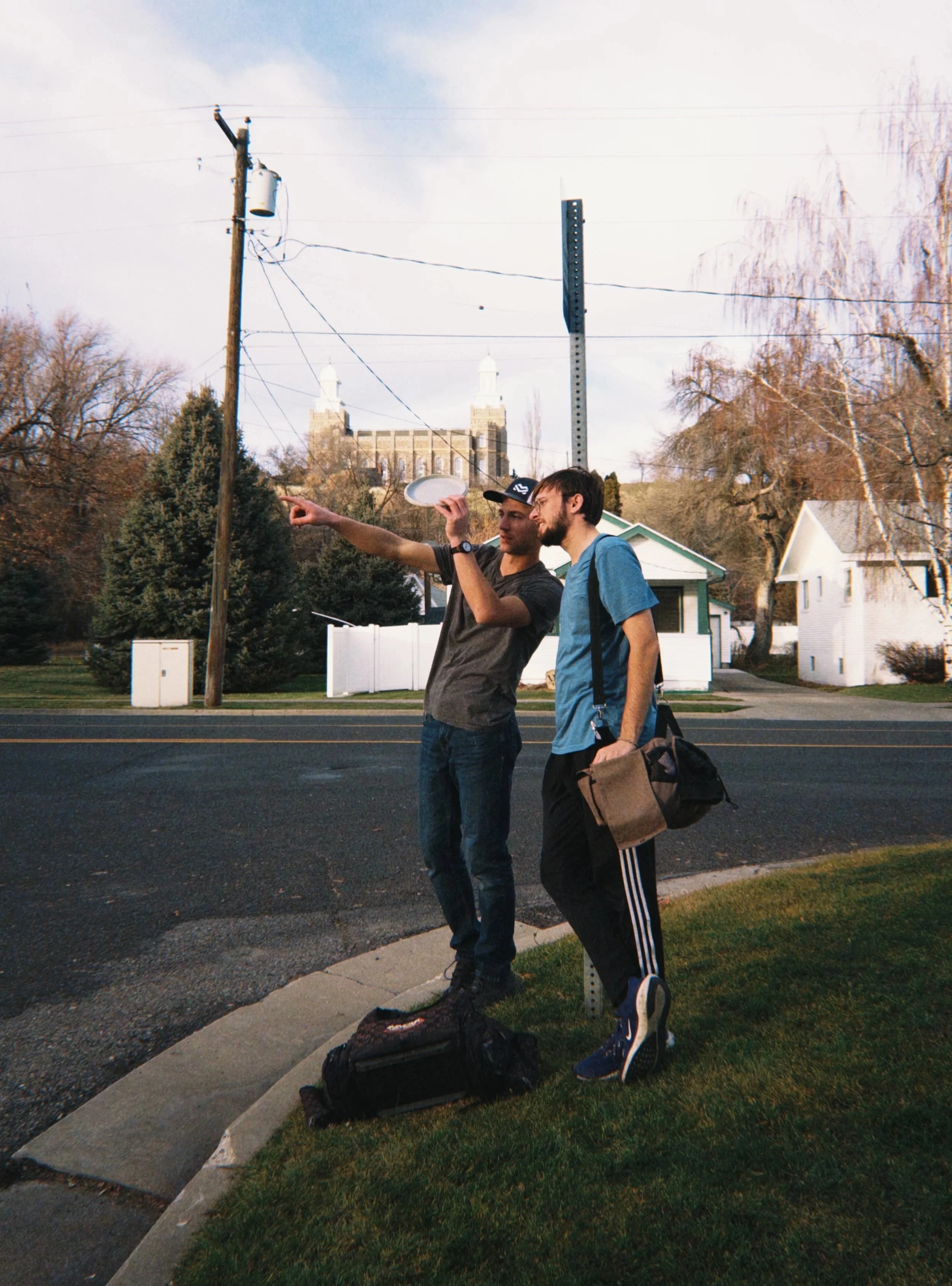 Two young men standing on a sidewalk near a street, with a white house, trees, and a church with towers in the background. One man is pointing at something, holding a frisbee, and the other is engaged in conversation. There are bags on the ground nearby.